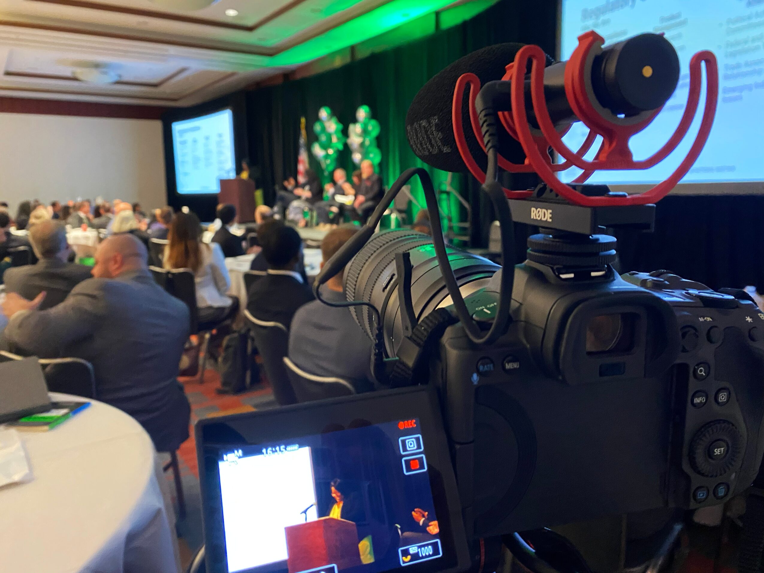Conference room with audience, presentation screen, and stage decorated with green plants, viewed through a camera's display.