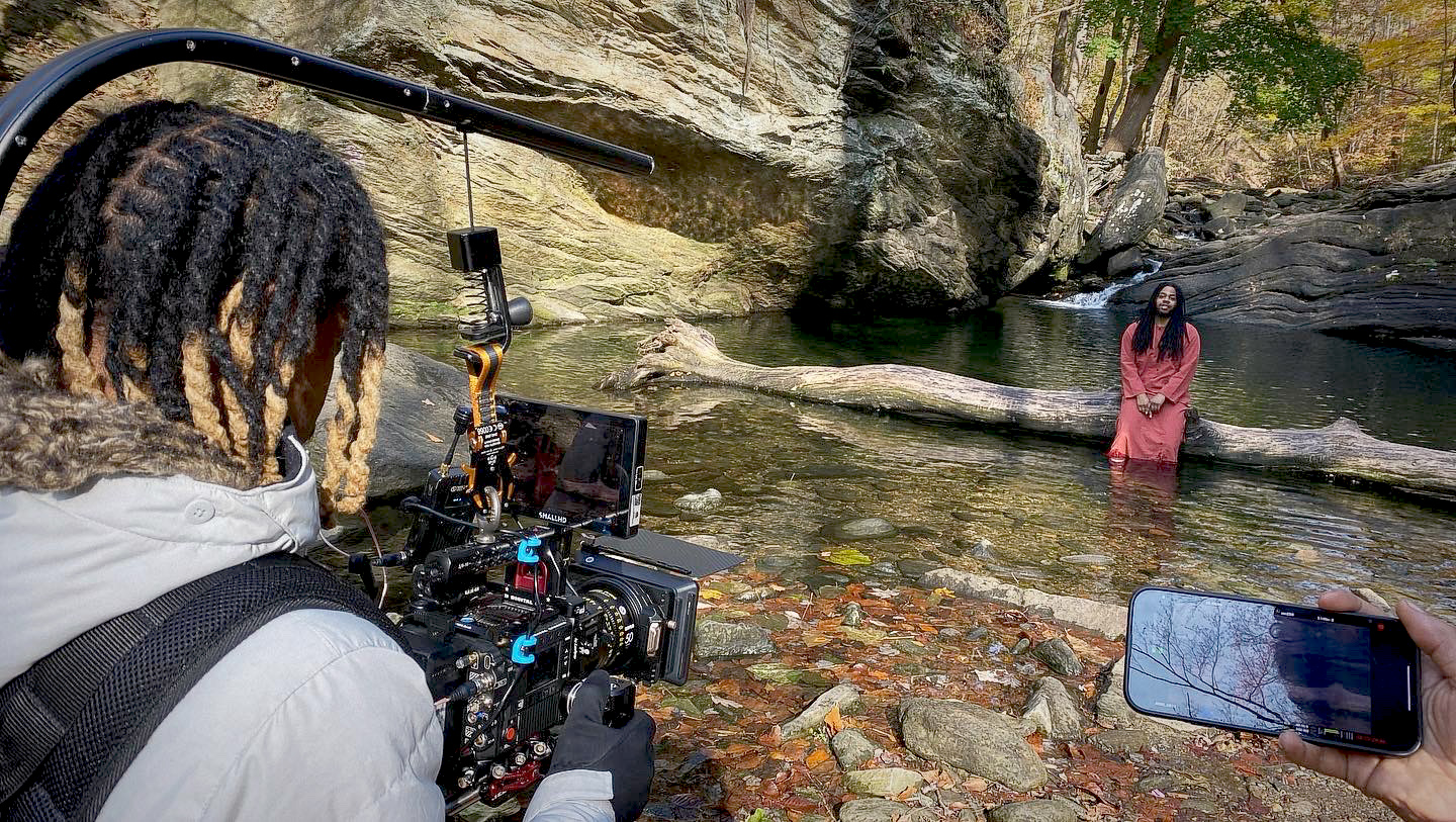 A person with dreadlocks films a woman standing in a river with rocks and trees in the background.