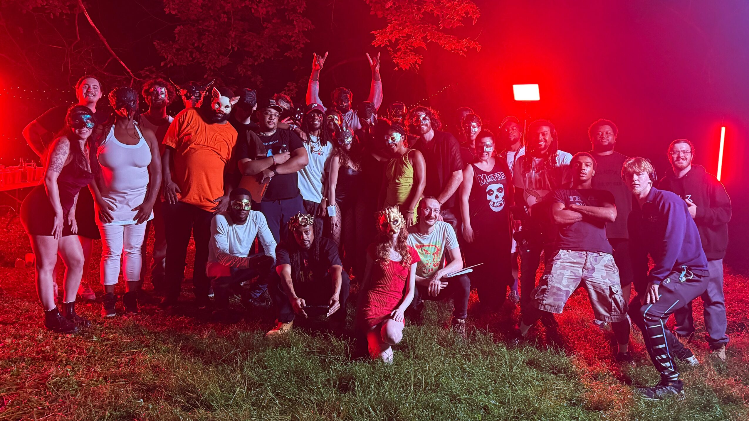 Group of people gathered outdoors at night, some standing and some sitting, with red and purple lighting effects.