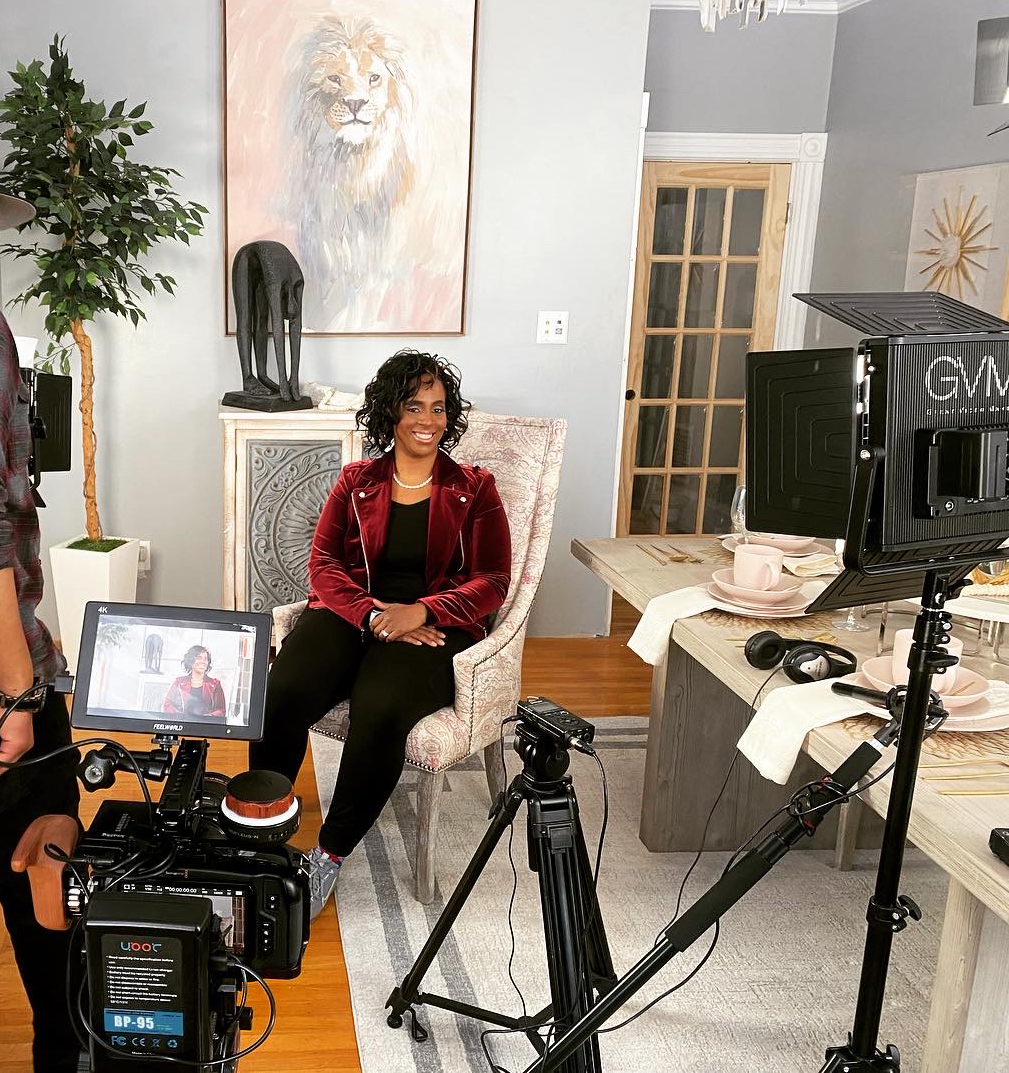 Woman with curly hair in red jacket sitting on a chair in a room with artwork and plants, being filmed or photographed.