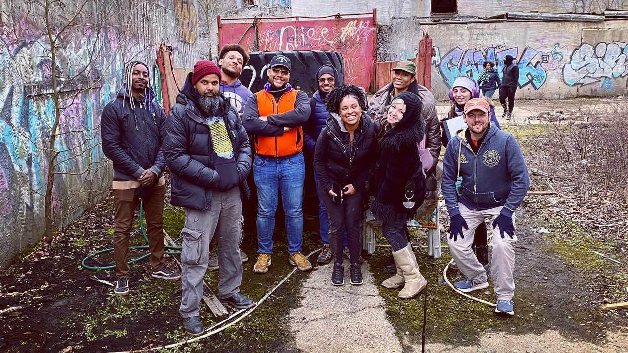 Group of people standing outdoors in front of graffiti-covered walls, some smiling and wearing jackets.