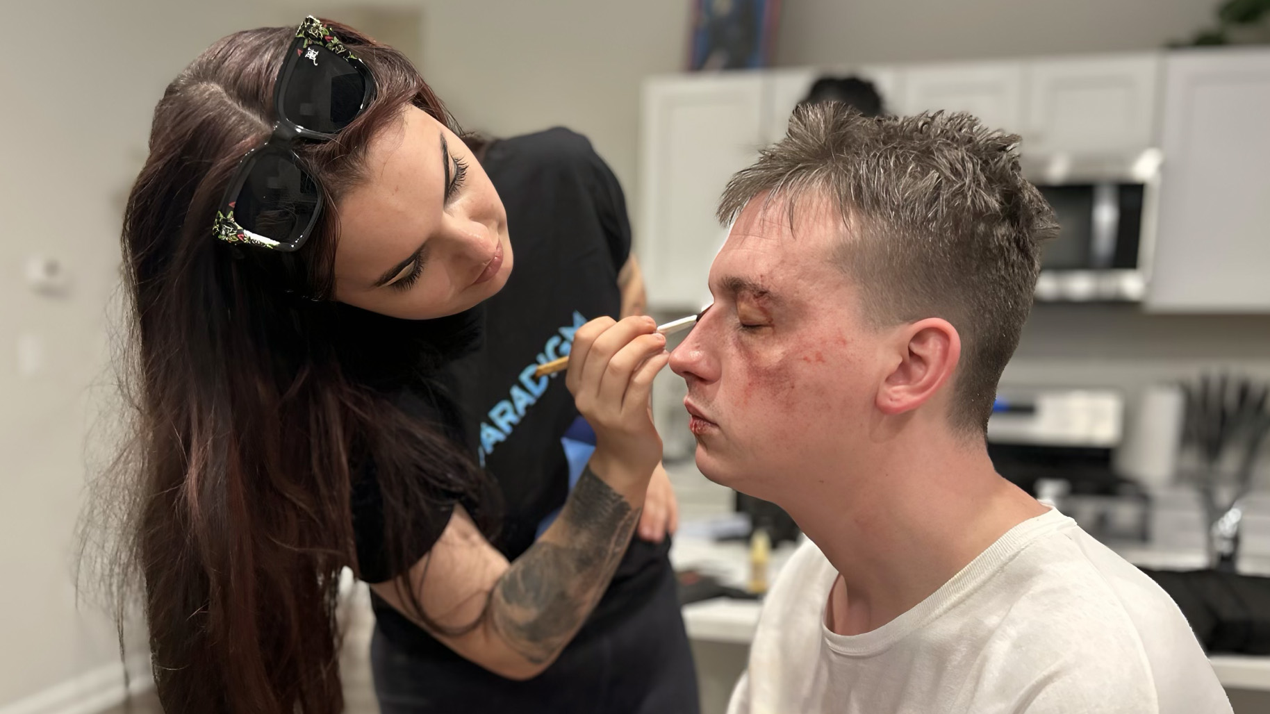 Woman applying makeup to a young man in a kitchen setting, with cabinets and appliances in the background.