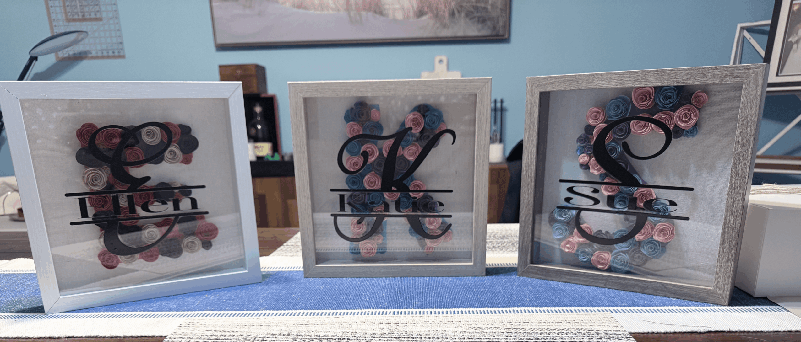 Three framed monogram art pieces with floral backgrounds, displayed on a table, with a blue wall and picture frame behind.
