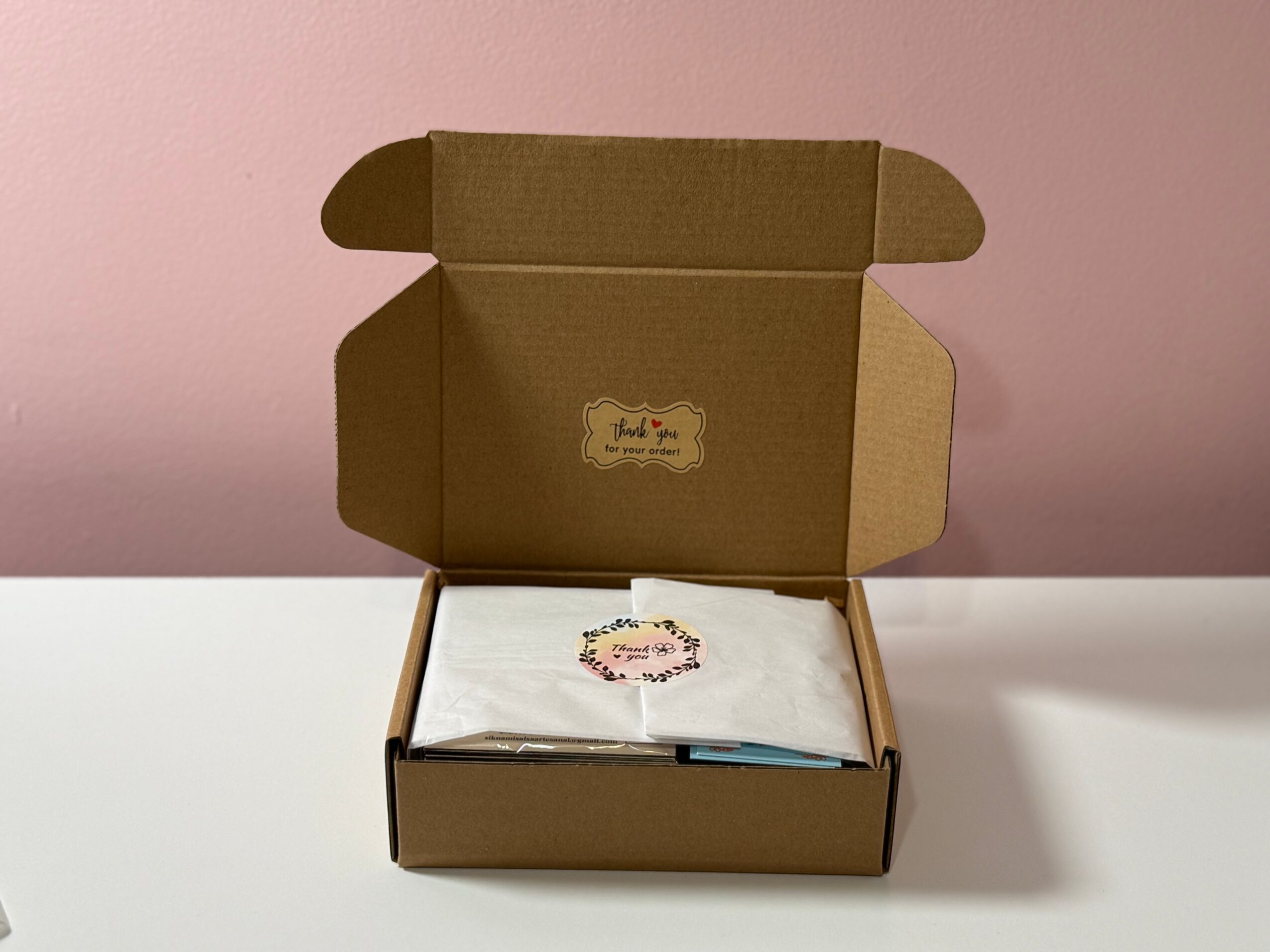 Open cardboard box with tissue paper inside on a white surface against a pink background.