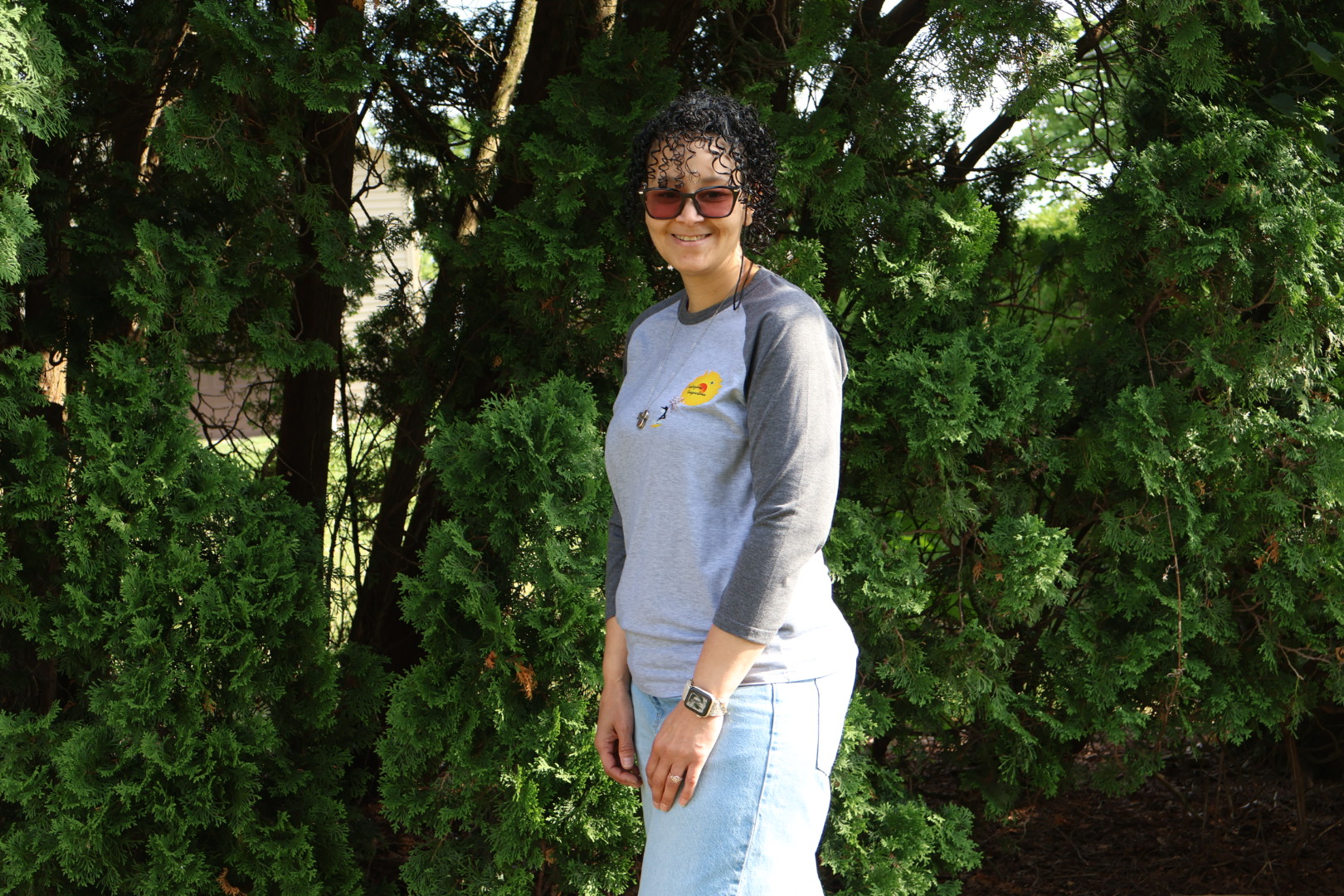 Woman with sunglasses and curly hair standing outdoors in front of green trees, smiling, wearing a gray long-sleeve shirt and white pants.
