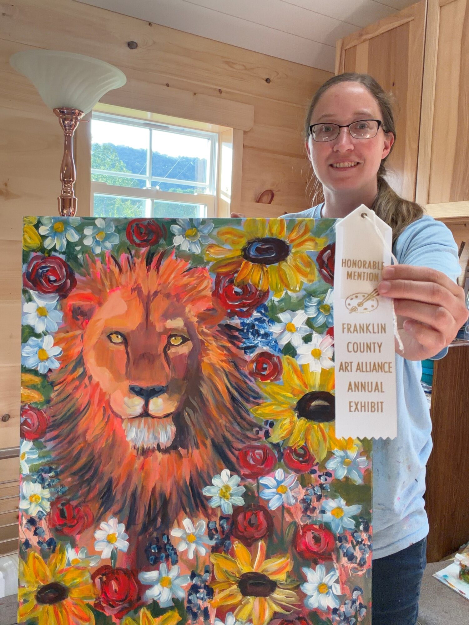 Woman holding colorful lion painting with a white tag, standing in a room with wooden walls and a window.