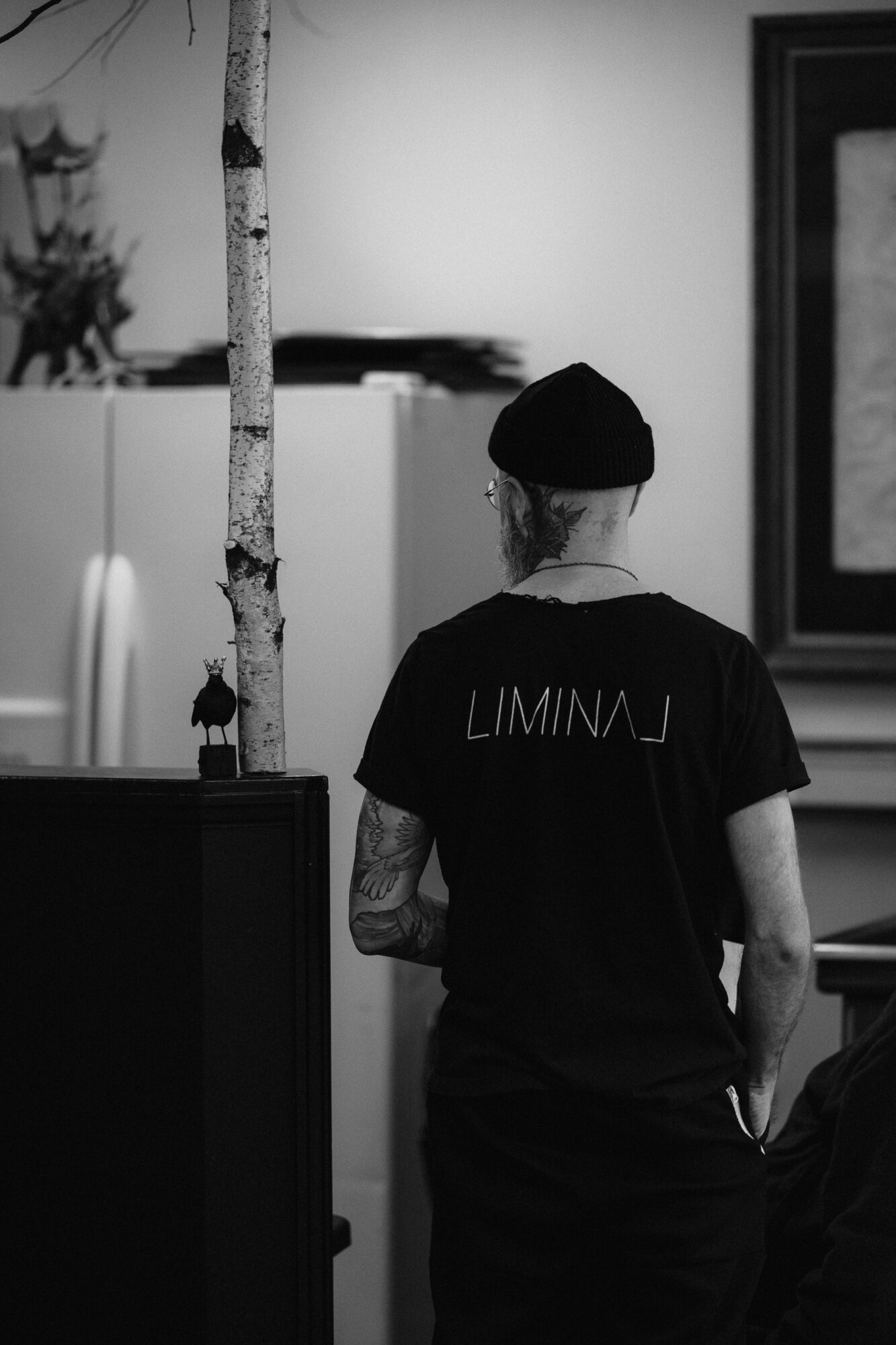 Person with a tattooed arm wearing a black beanie and T-shirt with text, standing indoors near a tree and framed artwork.
