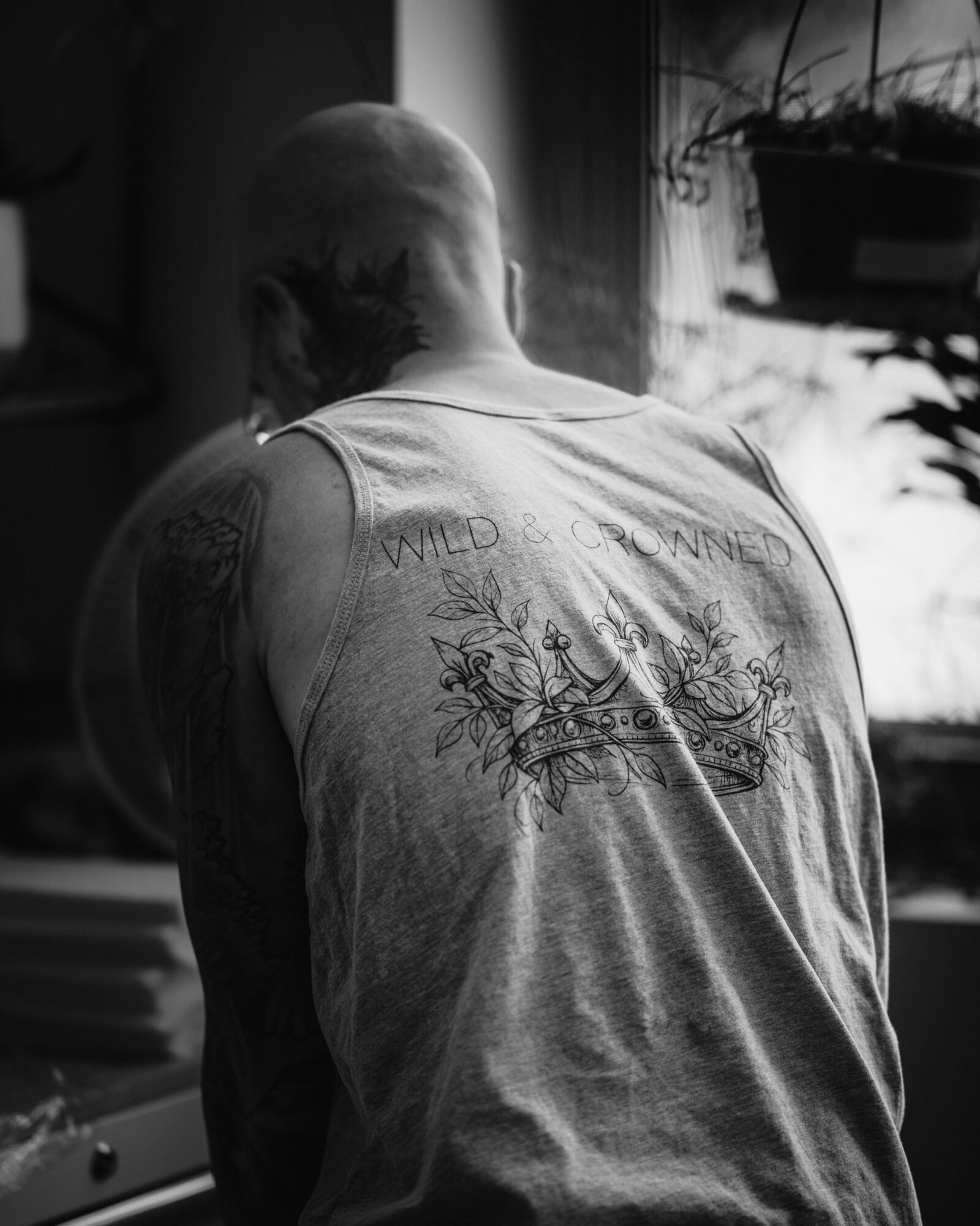Back of a person with a shaved head wearing a sleeveless shirt with a tattoo on the arm, indoors near a window.