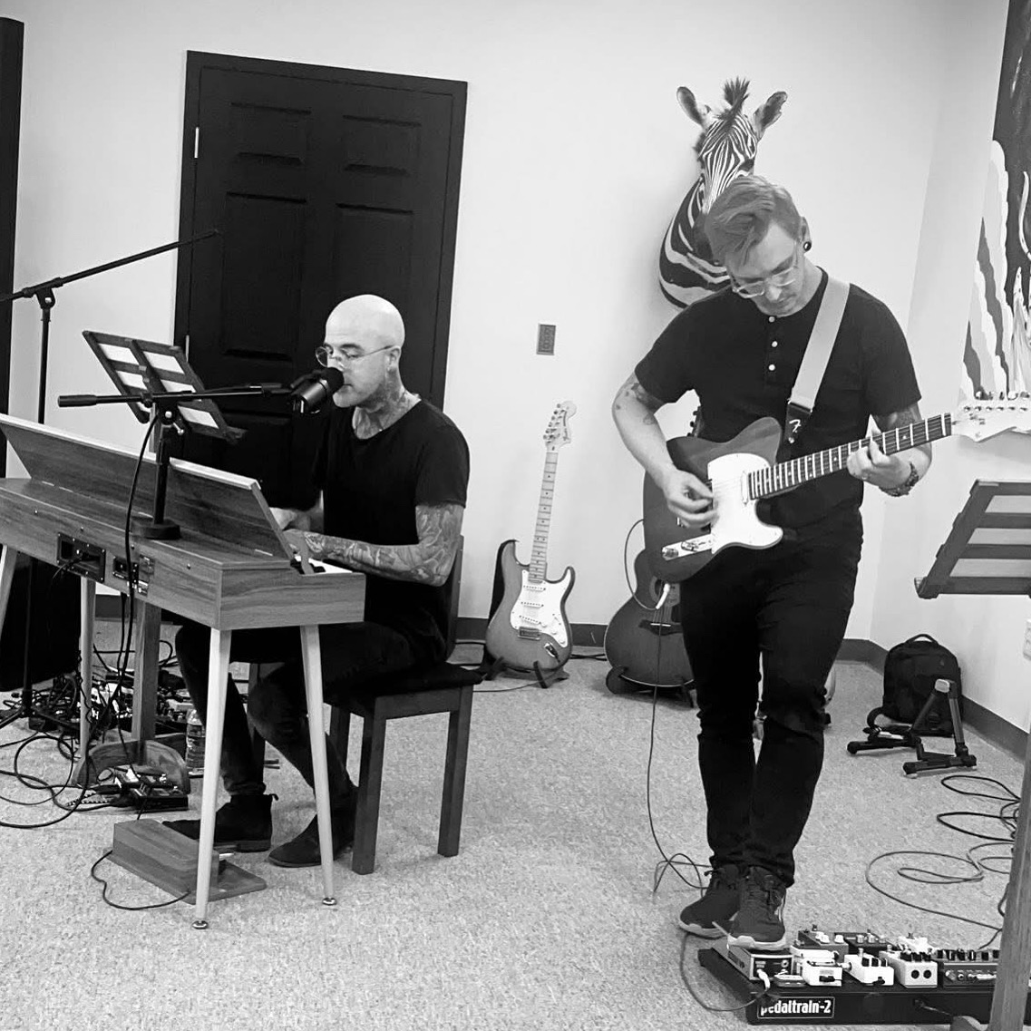 Two musicians perform indoors; one plays keyboard, the other guitar, with musical equipment and wall decorations visible.