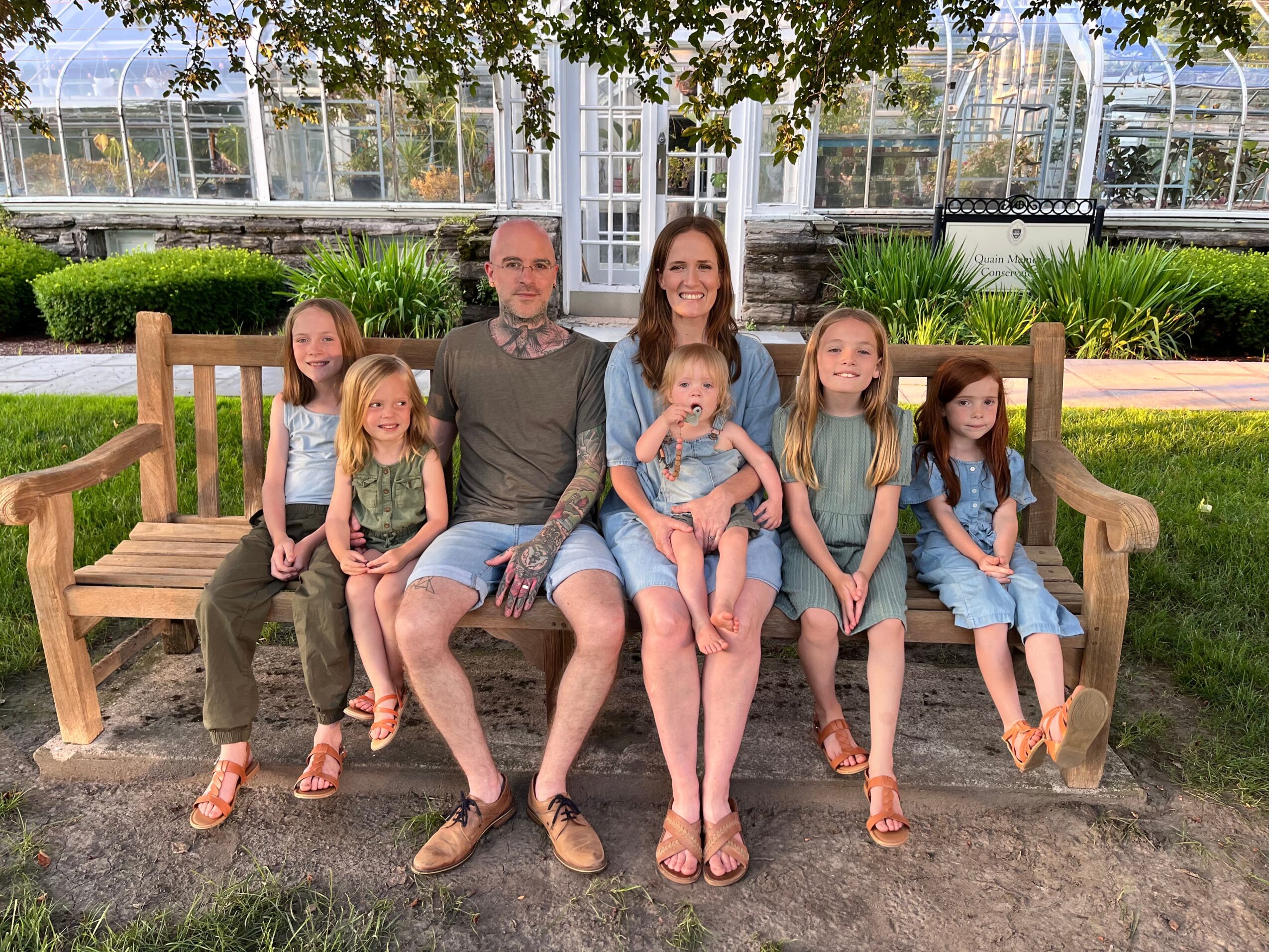 Family of seven sitting on a wooden bench outdoors, with children and two adults smiling, greenery, and a greenhouse in background.