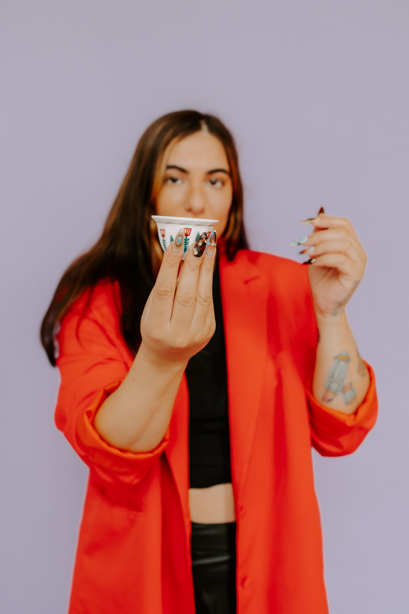 Woman with long hair in a red jacket holding a colorful cup, looking at the camera, against a light purple background.