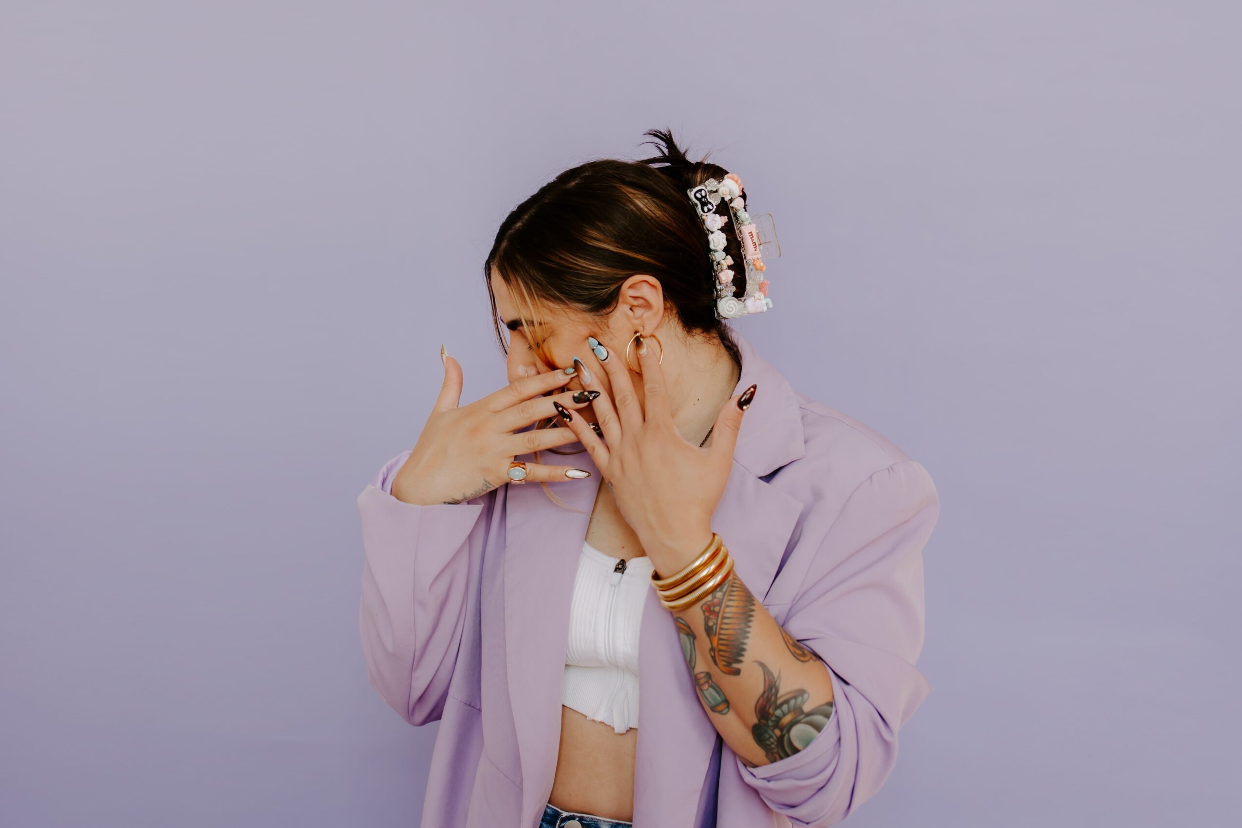 Woman with tattoos and jewelry covers her face with her hands, wearing a light purple jacket and white top.