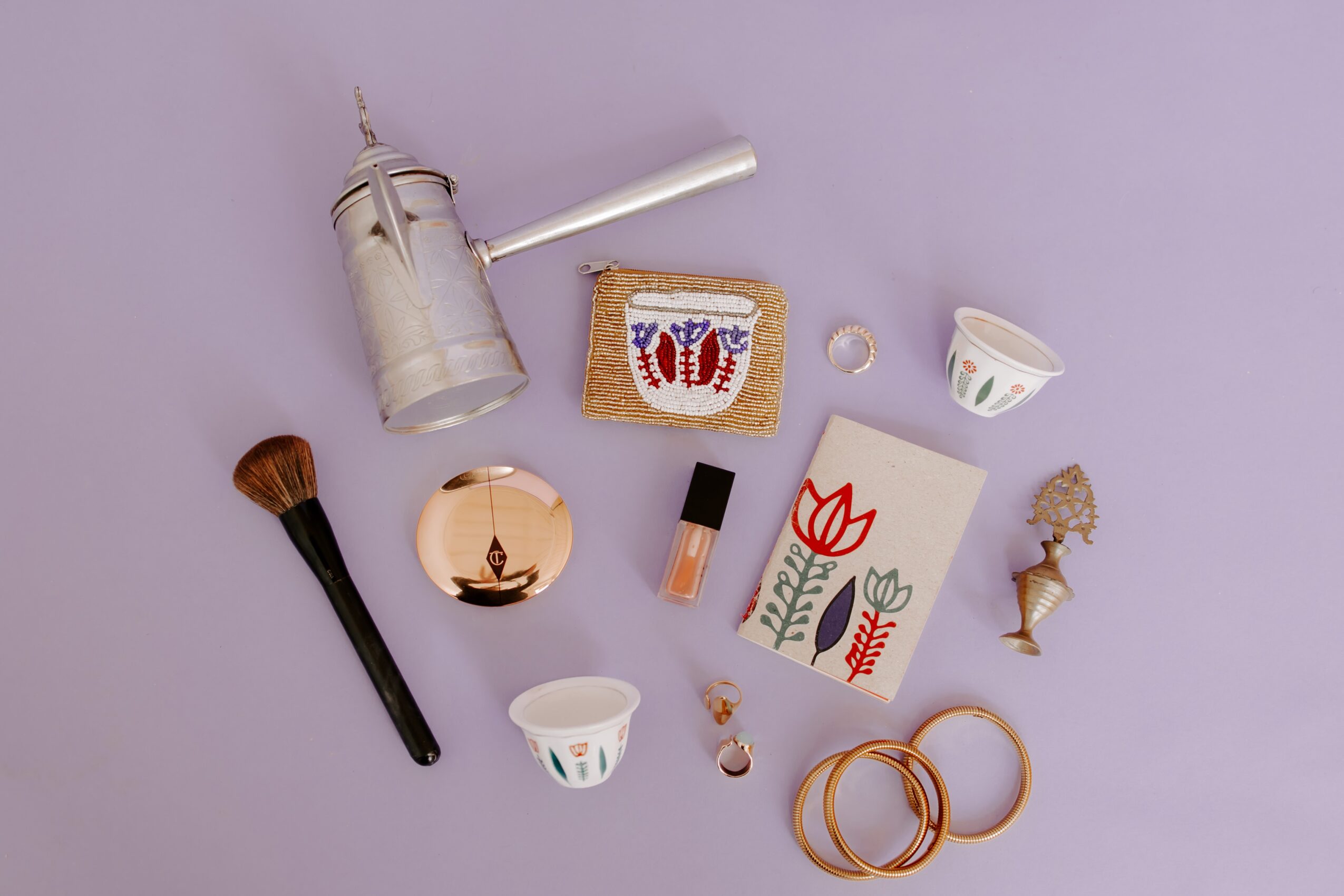 Flat lay of makeup and decorative items on a light purple background, including brushes, a candle, and artwork.