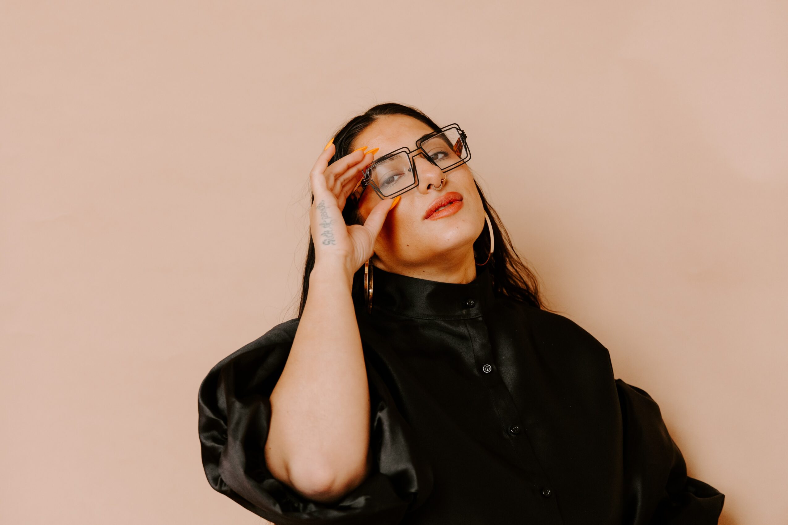 Woman with glasses and long hair, wearing a black top, touching her glasses, against a plain background.