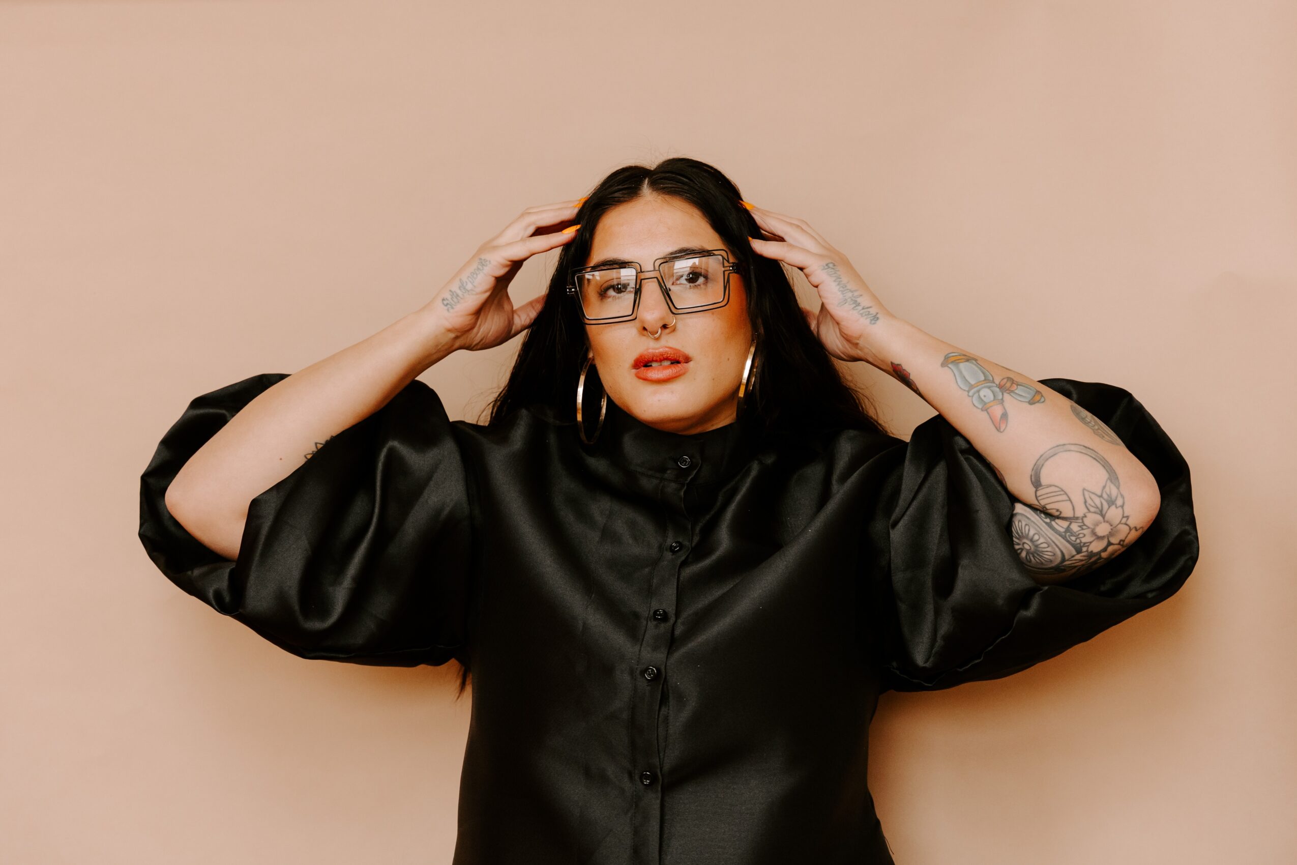 Woman with black hair, glasses, and tattoos adjusting her glasses against a beige background.