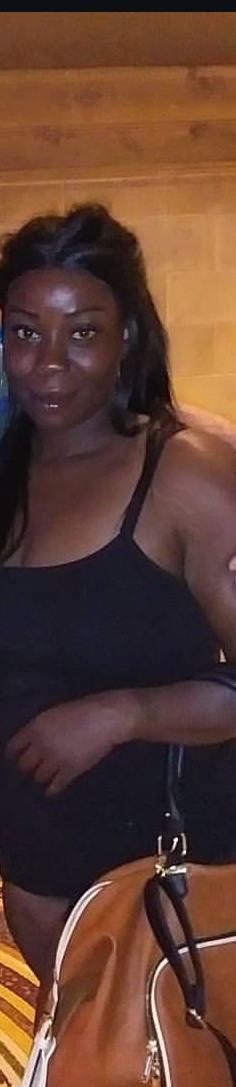 Woman with dark hair and a black top, holding a bag, standing indoors with wooden background.