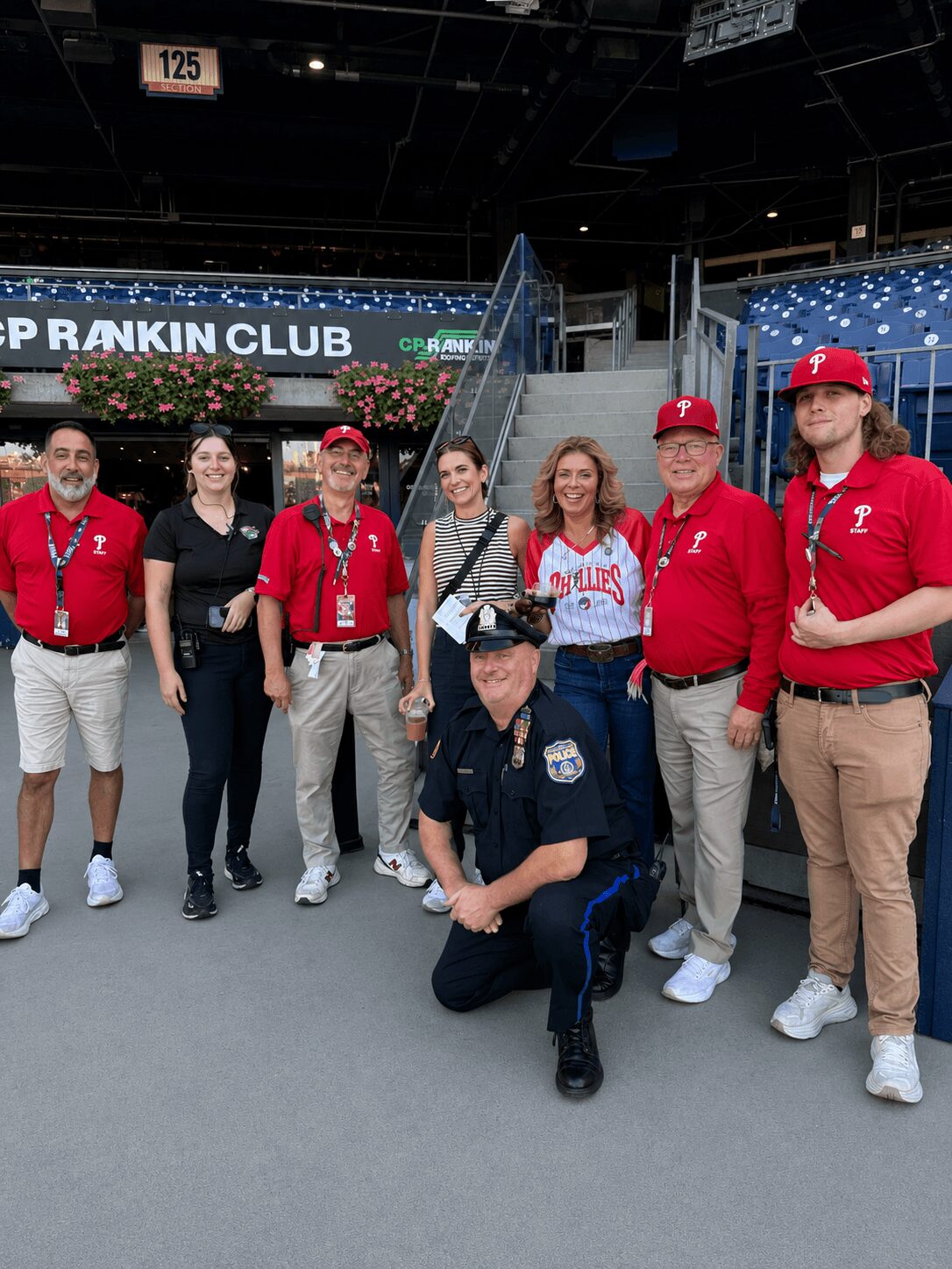 Group of nine people, including a police officer, standing in a stadium with a sign reading 'Top Rankin Club' in the background.
