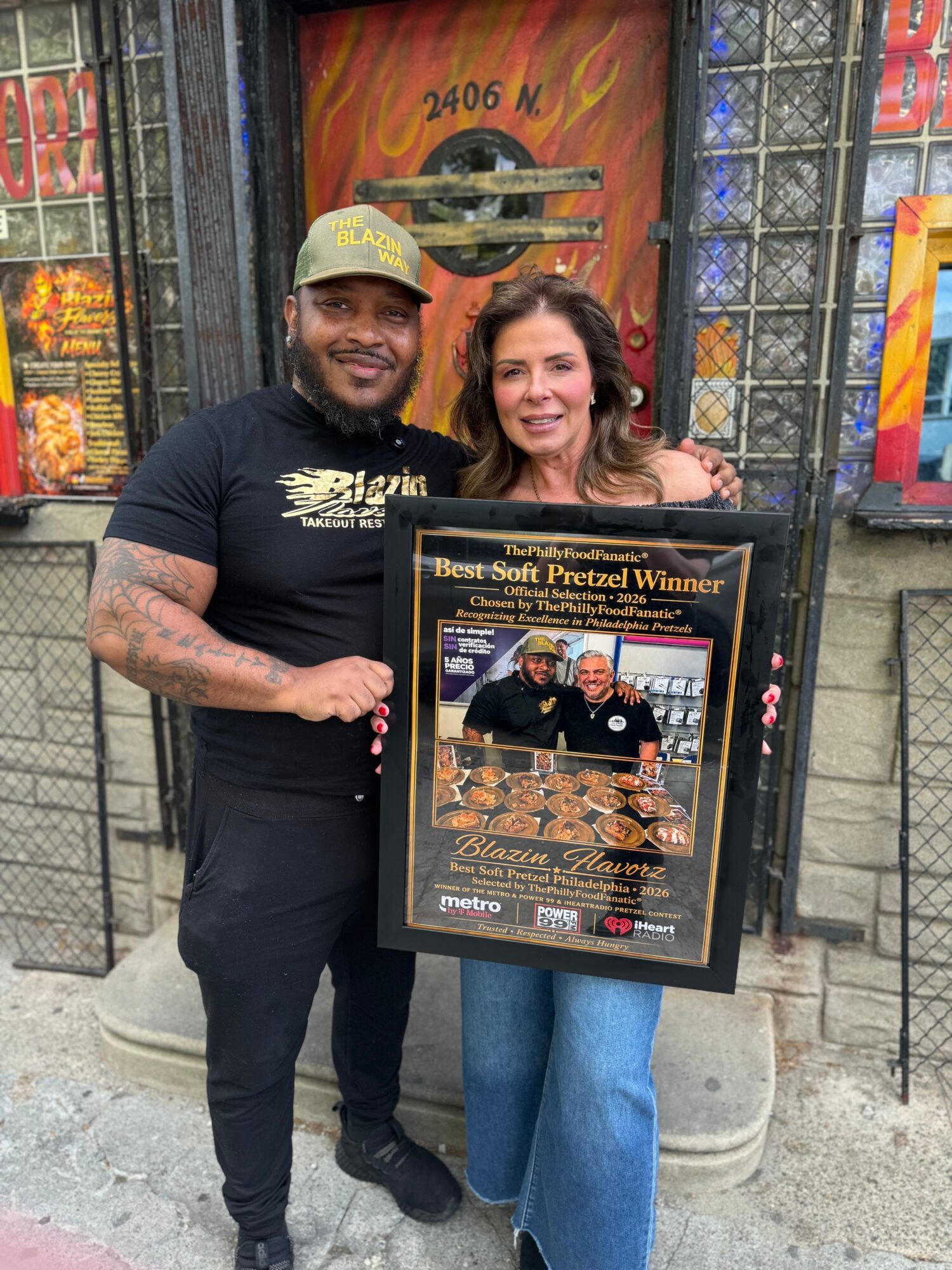 A man and woman stand outside a building holding a framed award for Best Soft Pretzel Winner, smiling at the camera.