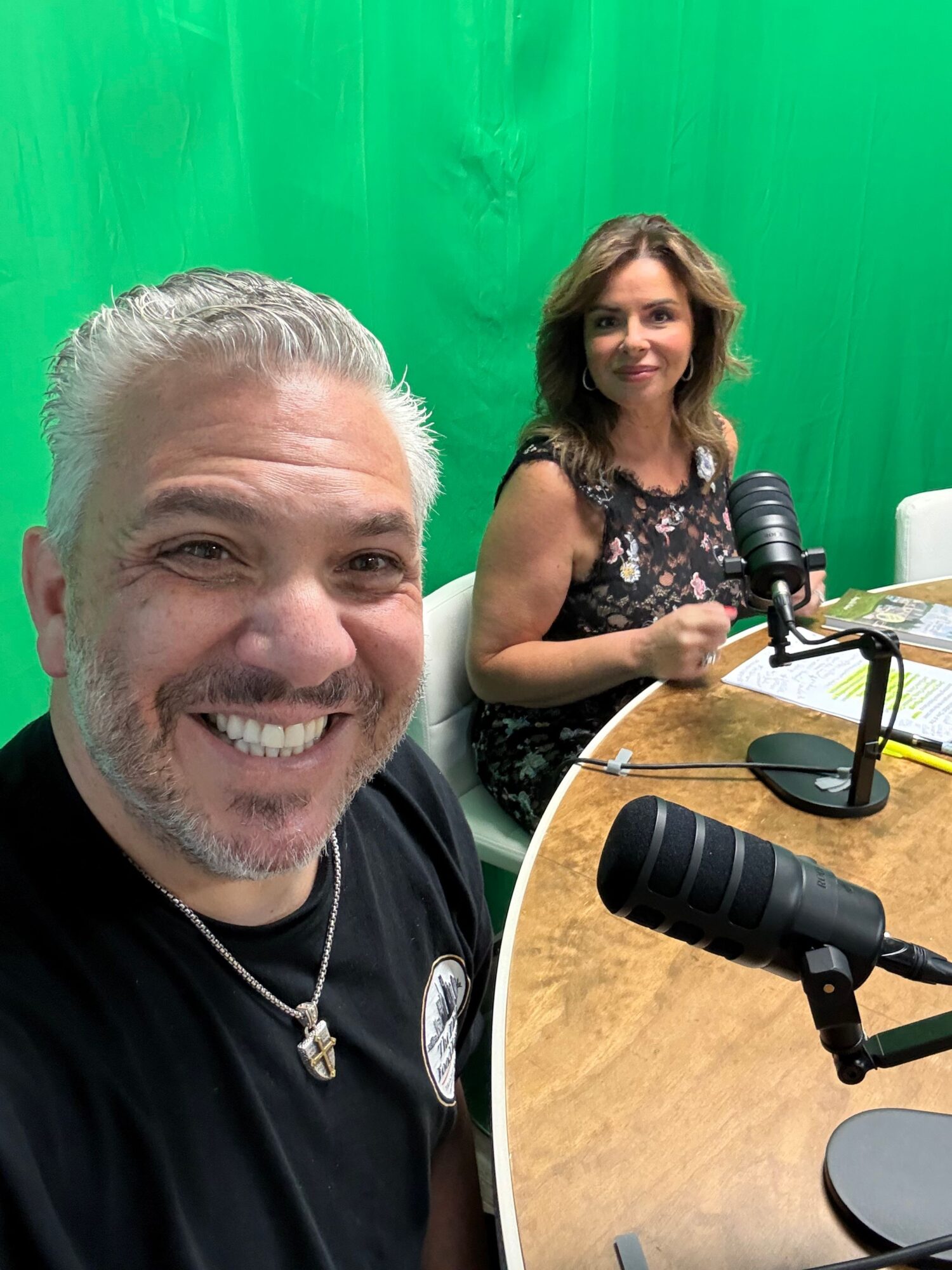 Two people in a studio with a green screen, one smiling at the camera, the other sitting at a table with a camera and microphone.