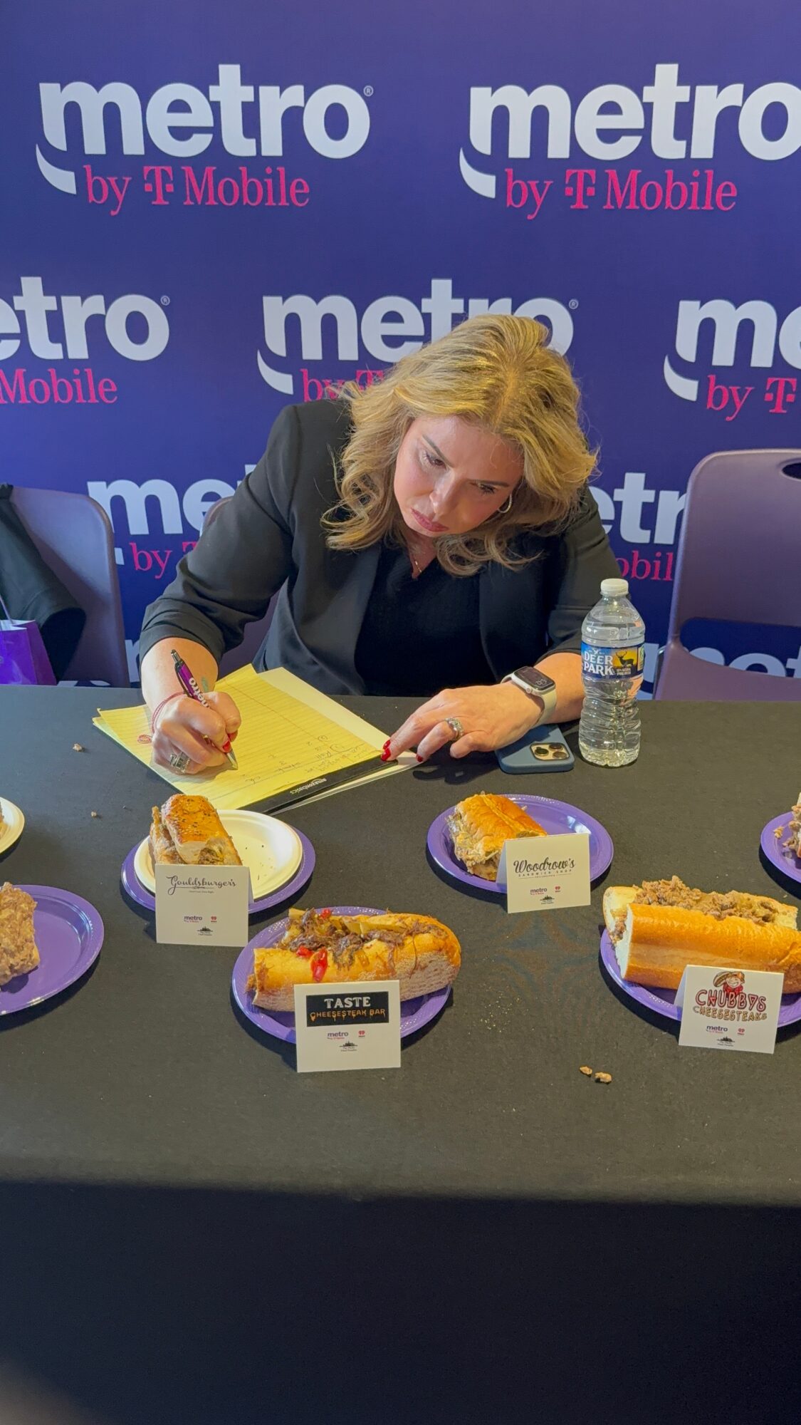 Woman signing papers at table with desserts and water bottle, backdrop with 'metro' logos.