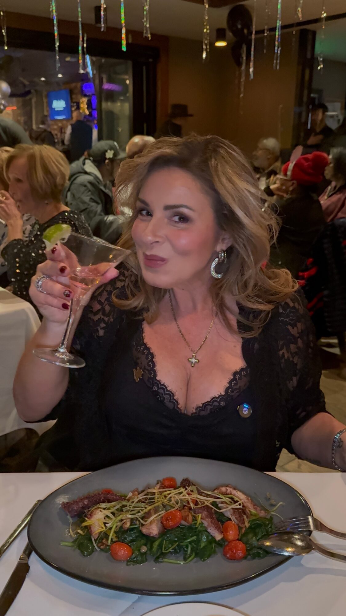 Woman with wavy hair holding a glass of wine at a restaurant, with a plate of pasta in front, surrounded by other diners.