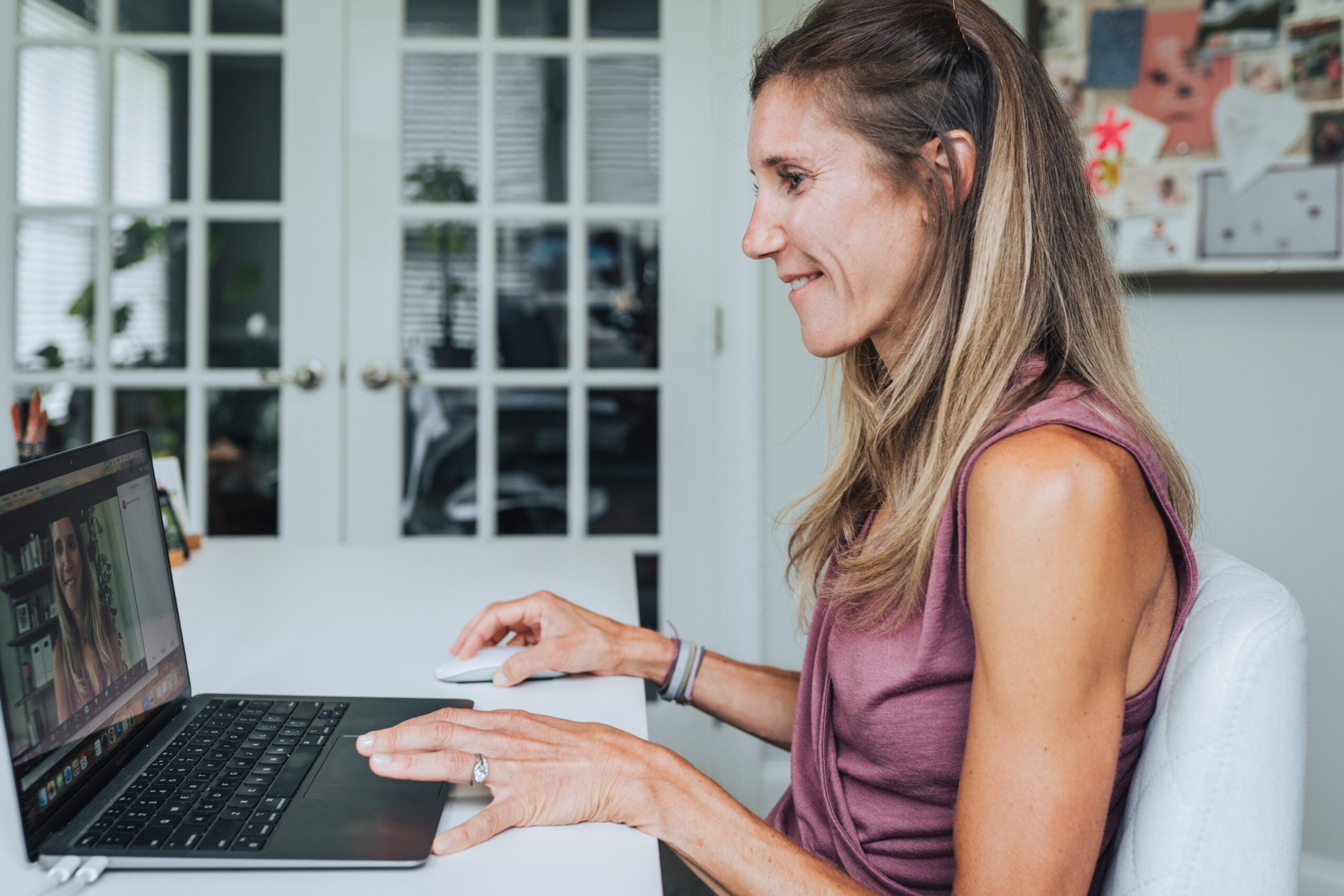 Woman sitting at a desk using a laptop, smiling, with a glass door and bulletin board in the background.