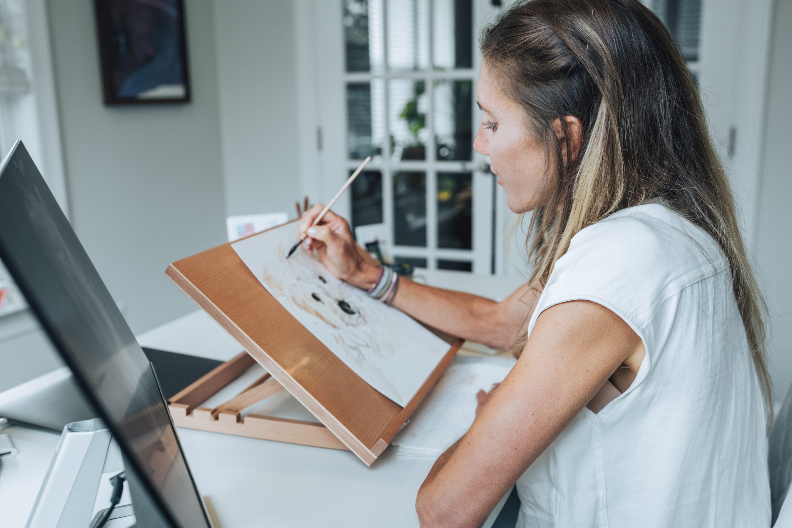Woman drawing on a sketchpad at a desk in a bright room with a window and door in the background.