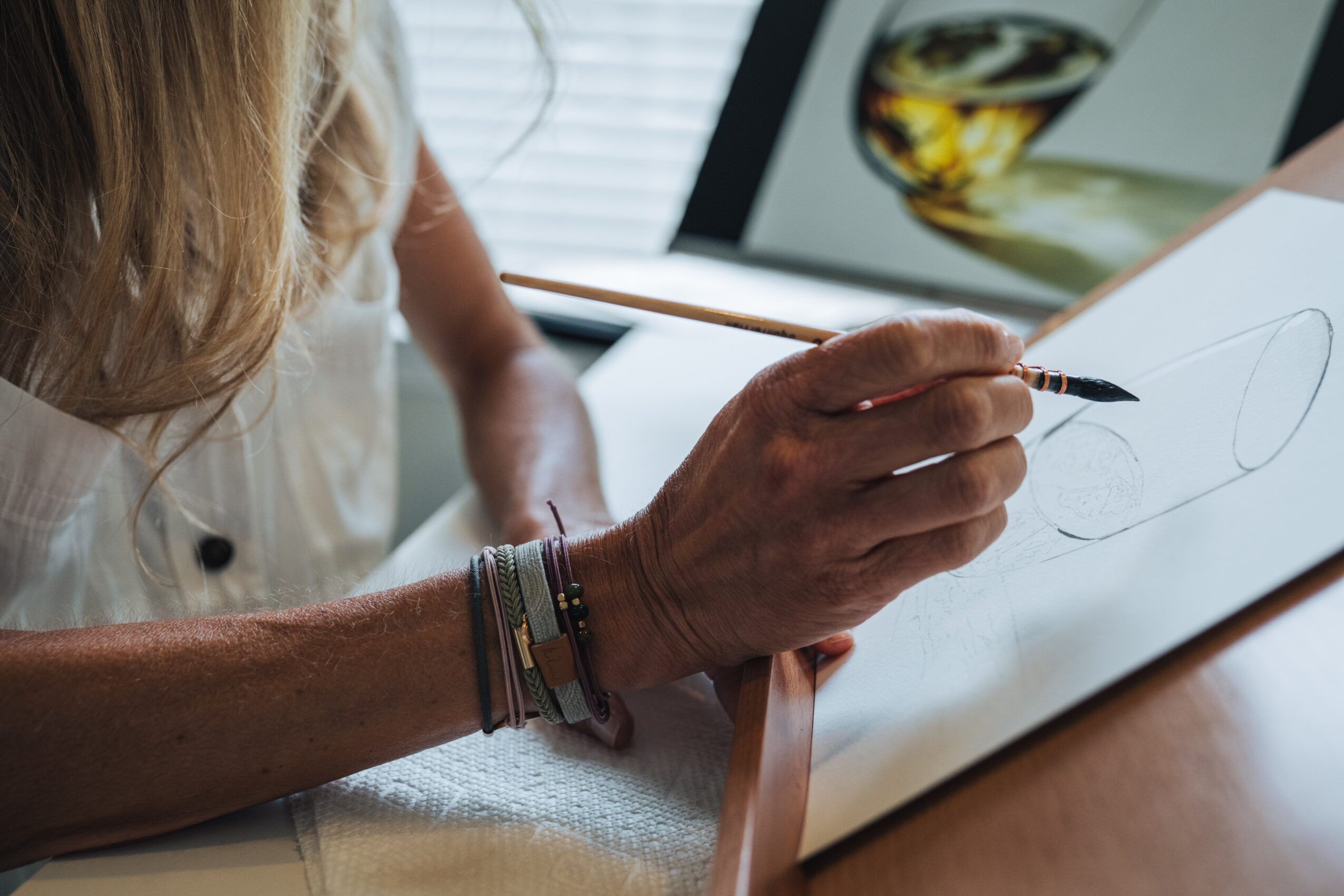 Person drawing on paper with a pen, with a tablet displaying a glass of wine in the background.