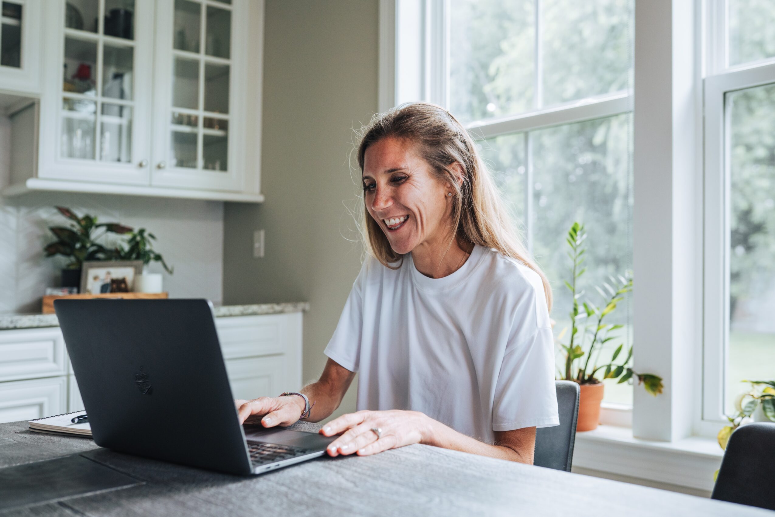 Woman smiling at a laptop in a bright kitchen with large windows and potted plants