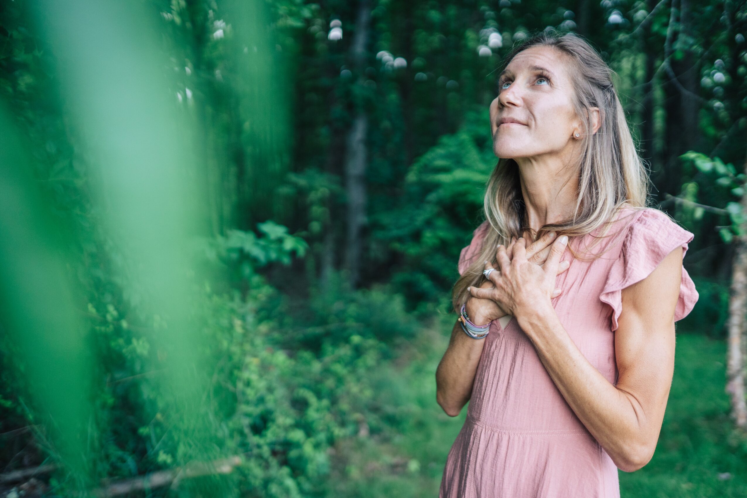 Woman in pink dress standing outdoors with hands on chest, looking upward, surrounded by green trees and foliage.