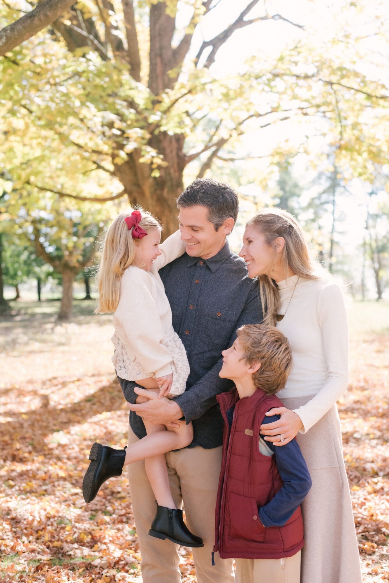 Family of four outdoors in autumn, with trees and sunlight, a man holding a young girl, woman and boy nearby