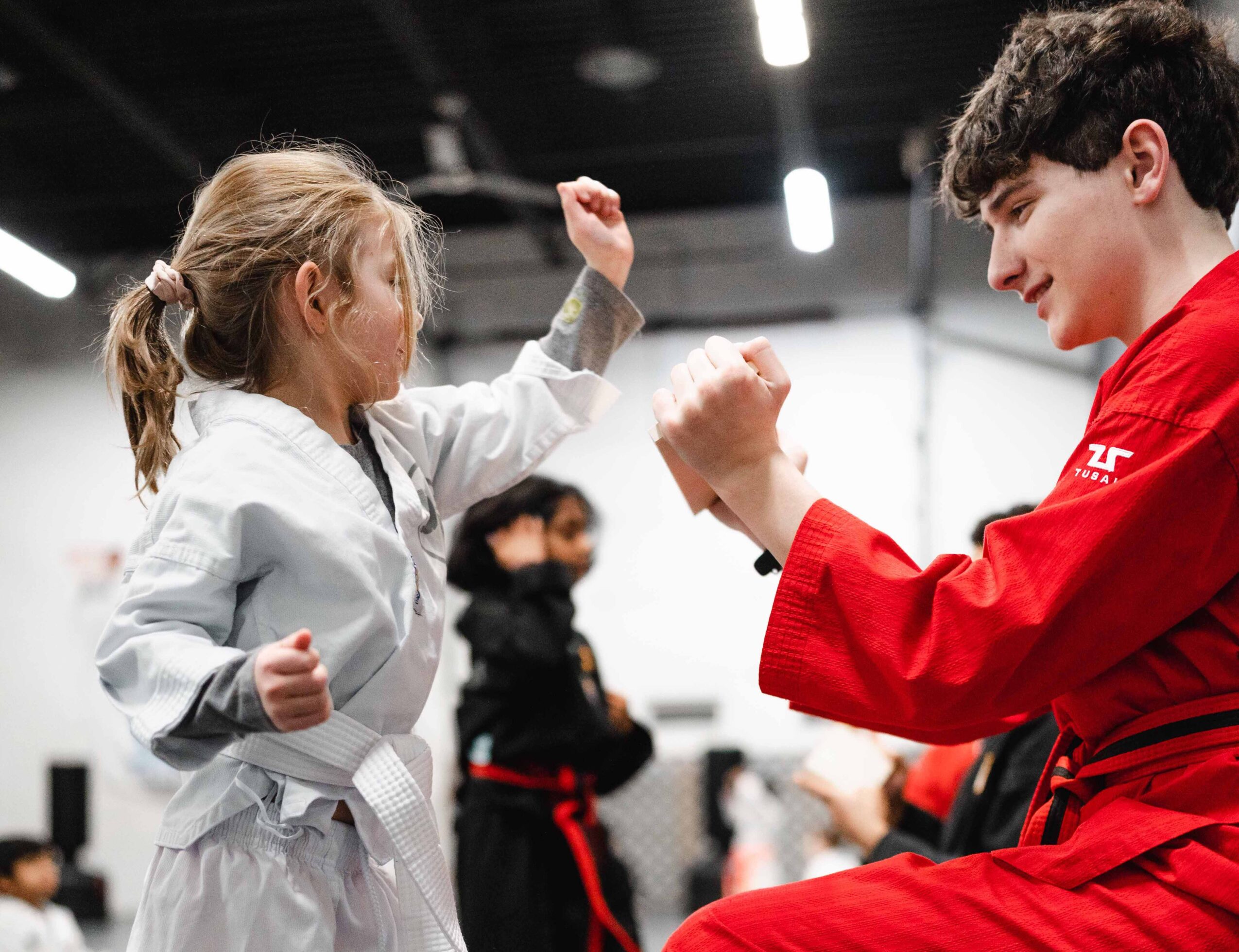 Two children in martial arts uniforms facing each other, practicing a move in a dojo or gym, with other students in the background.