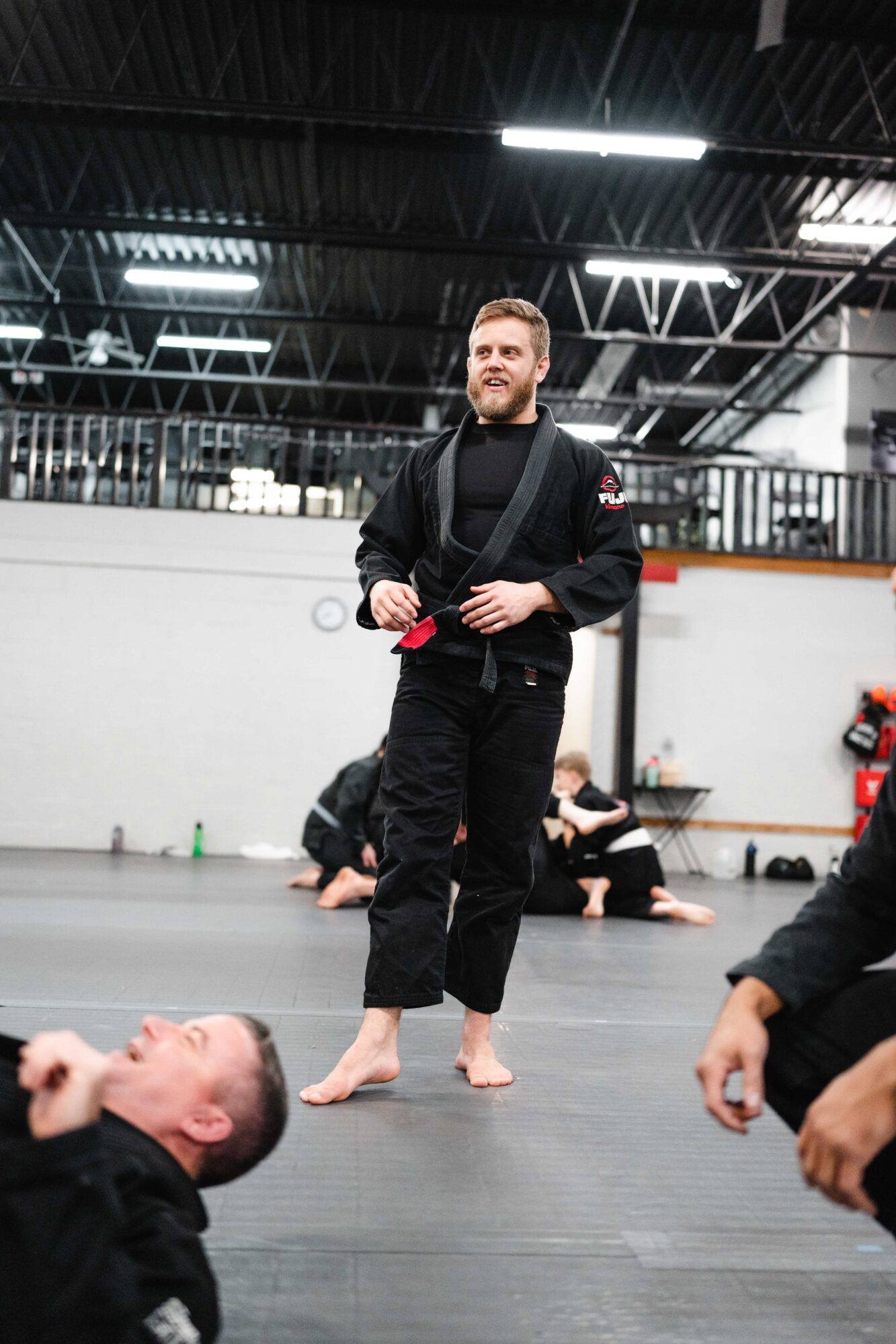 Man standing in a martial arts training facility, wearing a black gi, with people sitting on the floor in the background.