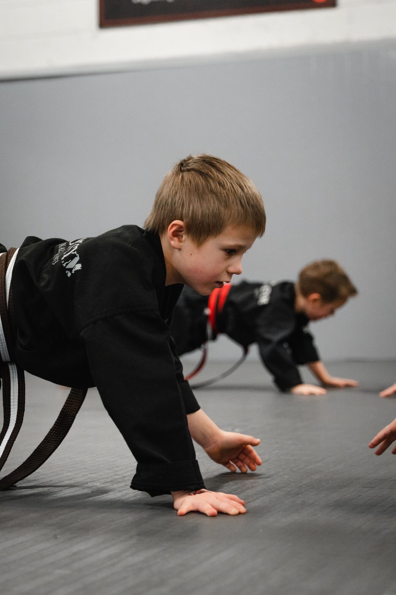 Two children in martial arts uniforms practicing crawling on a mat, focused and engaged.