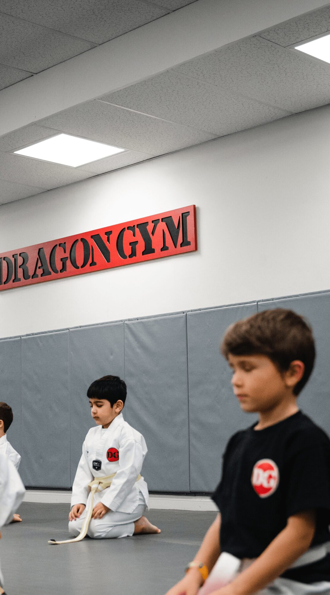 Children in martial arts uniforms sitting on the floor in a gym with a sign reading 'DRAGON GYM' on the wall.