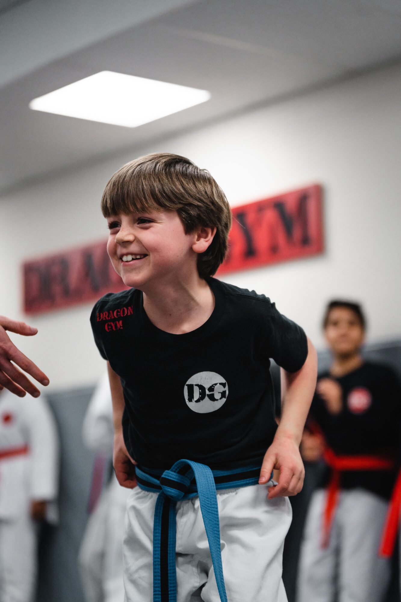 Smiling boy in martial arts uniform with a blue belt, in a training room with other children, smiling and looking to the side.