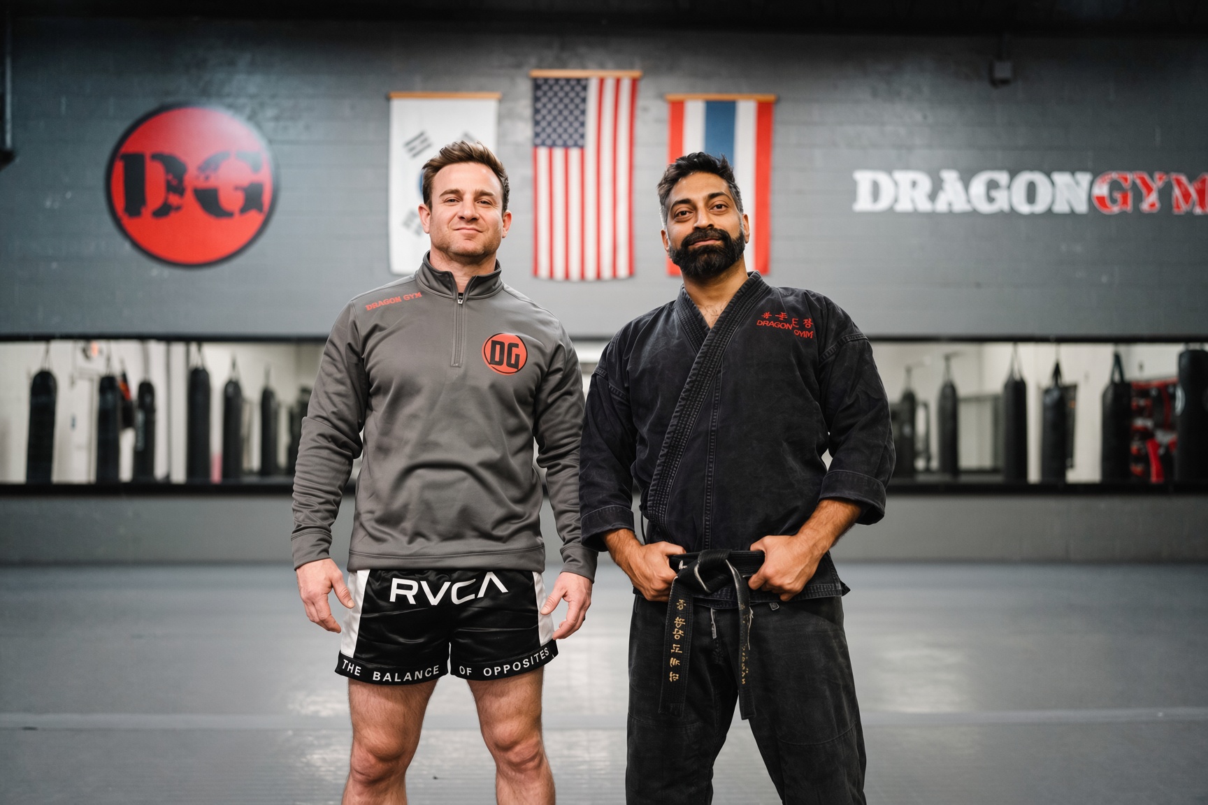 Two men stand in a gym with American and other flags behind them, one in a gray jacket and shorts, the other in a black gi, smiling.