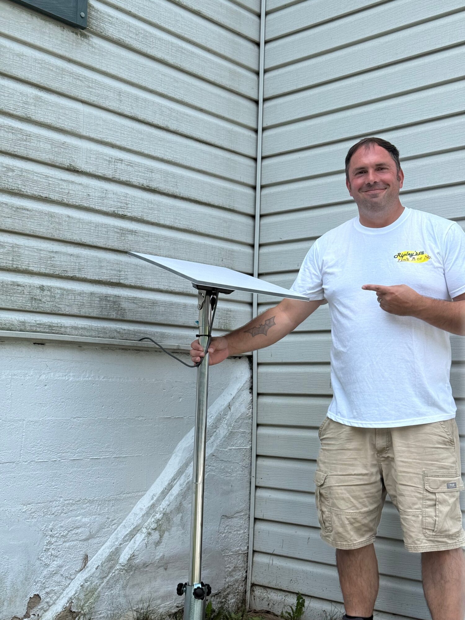 Man standing outdoors next to a metal pole, leaning on a white umbrella, wearing a white t-shirt and beige shorts.