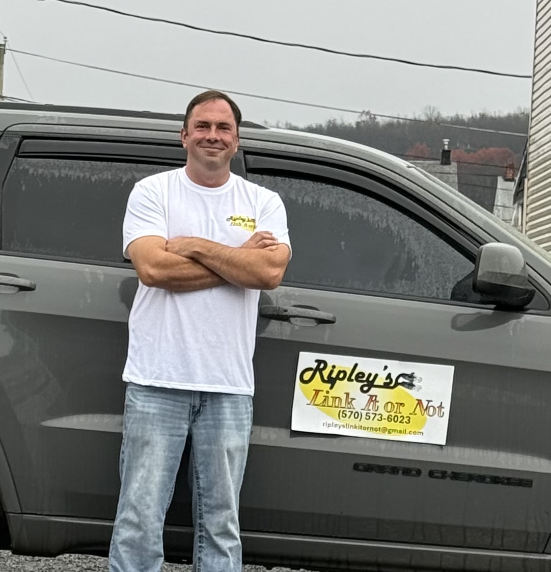Man standing with arms crossed next to a gray vehicle with a sign on the door, outdoors on a cloudy day.