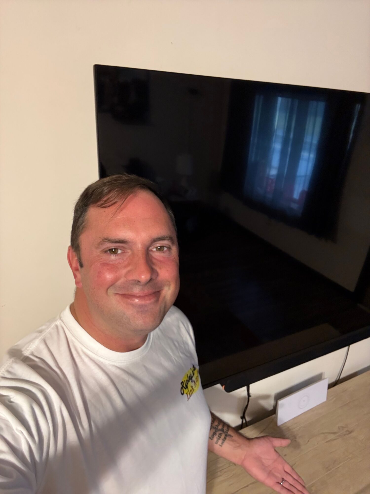 Person with short hair and a white shirt smiling, sitting at a table with a TV behind them.