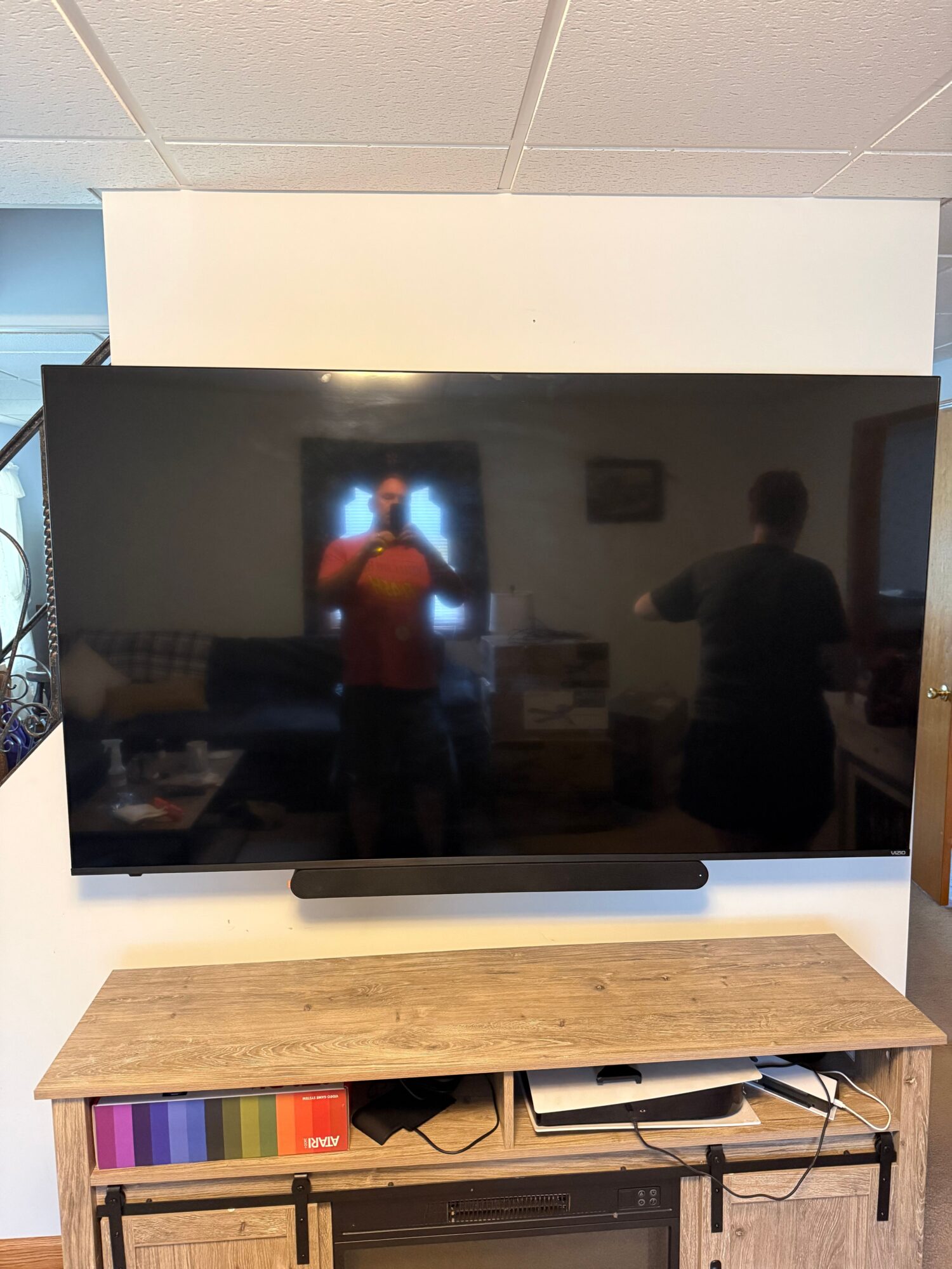 Flat-screen TV on a white stand with a wooden shelf, reflection of a person in a red shirt, and a colorful book on the side.