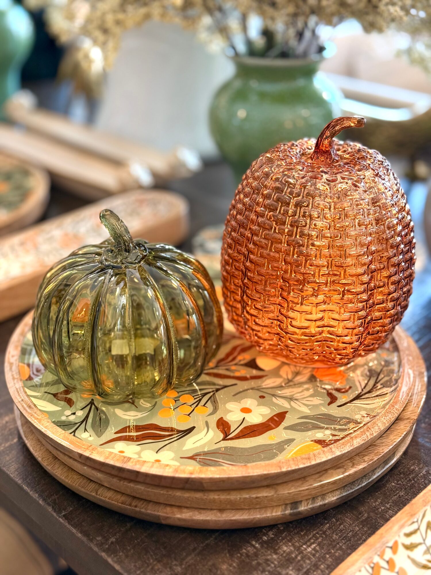 Two decorative glass pumpkins, one green and one amber, on a round tray with a floral pattern, in a cozy setting.