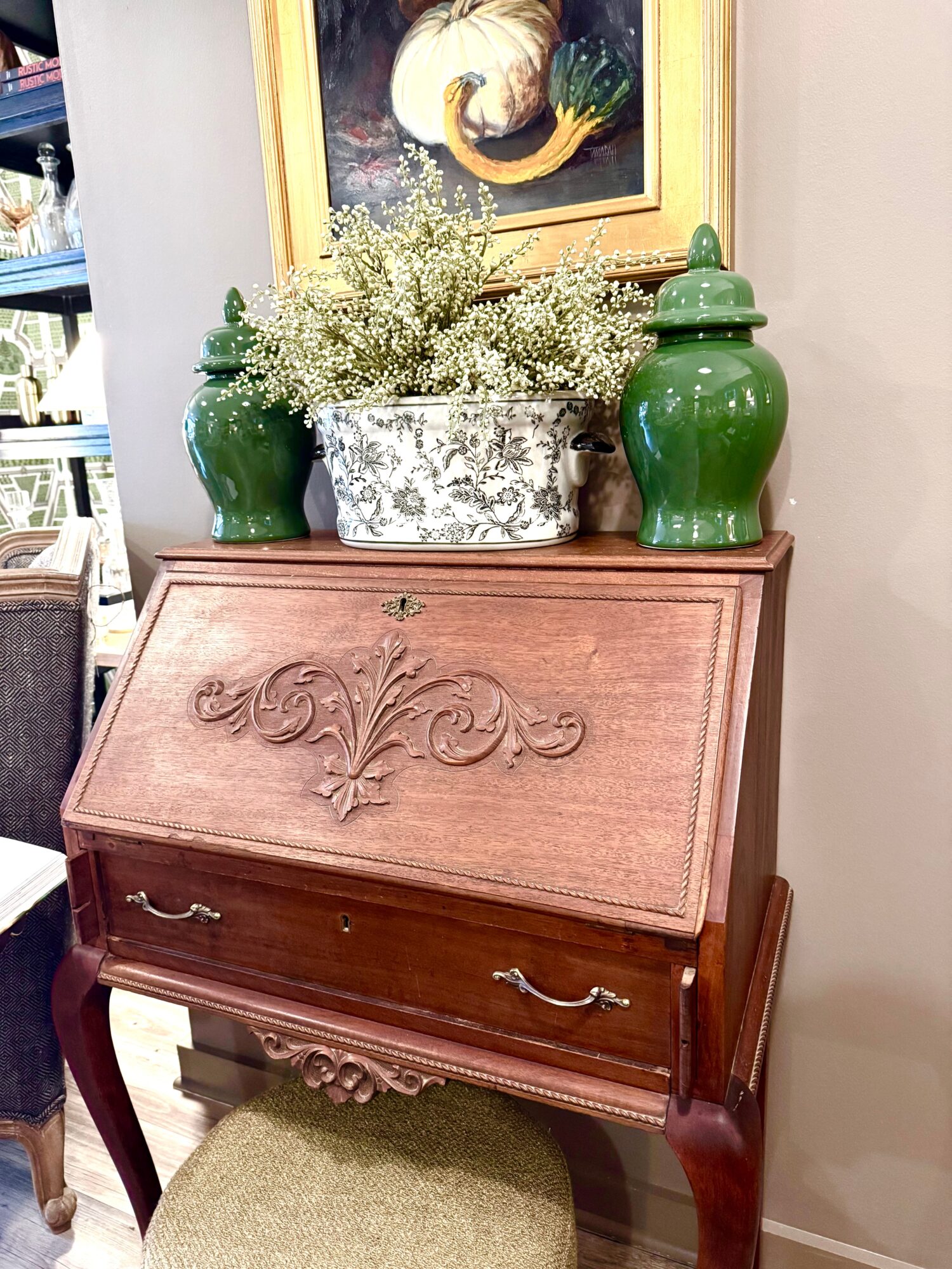 Wooden secretary desk with carved floral design, two drawers with metal handles, topped with green vases and a flower arrangement, framed artwork above.
