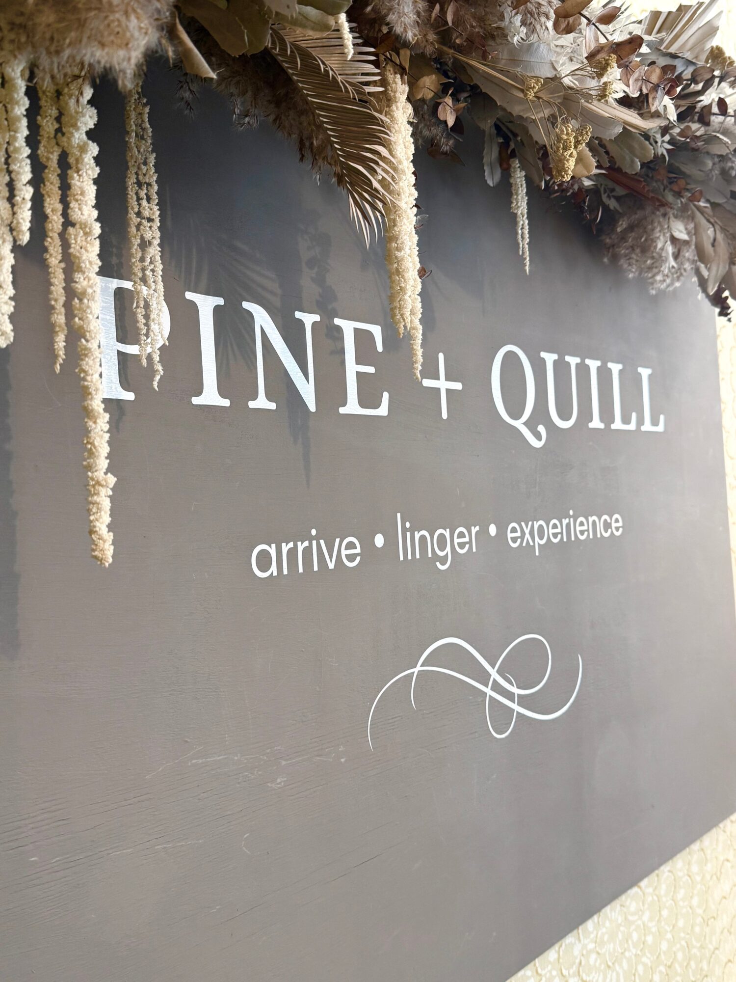 Sign for FINE + QUILL with text 'arrive linger experience' and decorative floral arrangement above.
