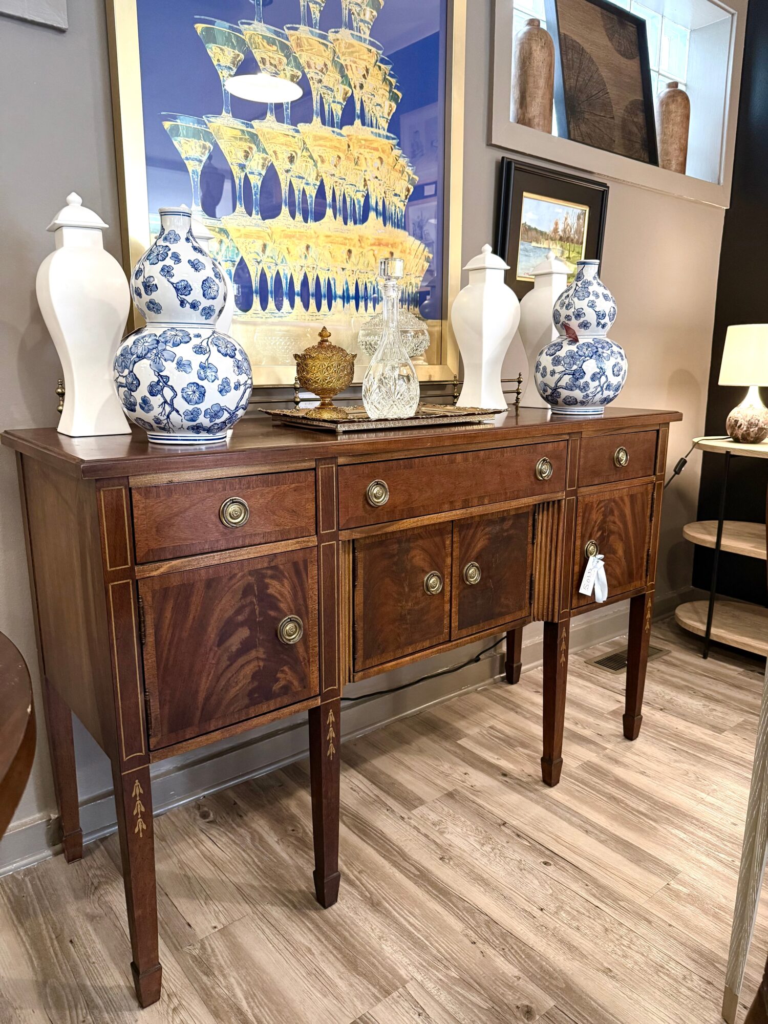 Wooden sideboard with decorative vases and artwork above, in a room with framed pictures and a lamp, on a wooden floor.