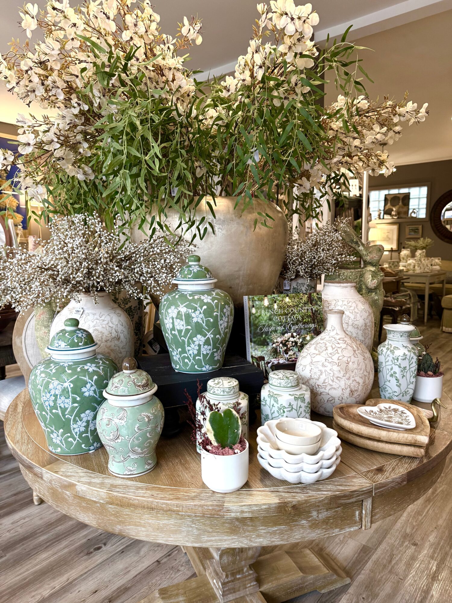 Arrangement of green and white vases with flowers on a wooden table, with a large floral display in the background.