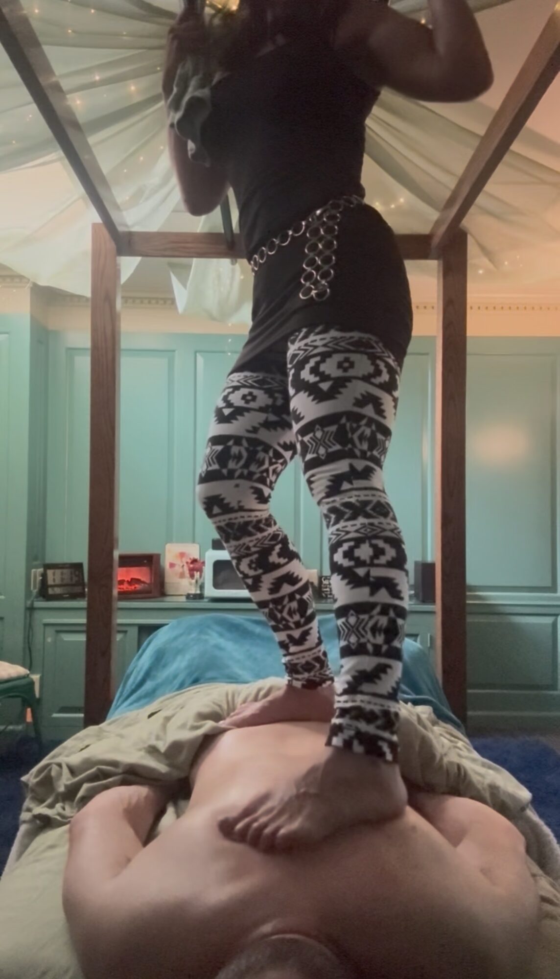 Person standing on another person's stomach in a room with teal walls and a mirror, wearing patterned pants and a black top.