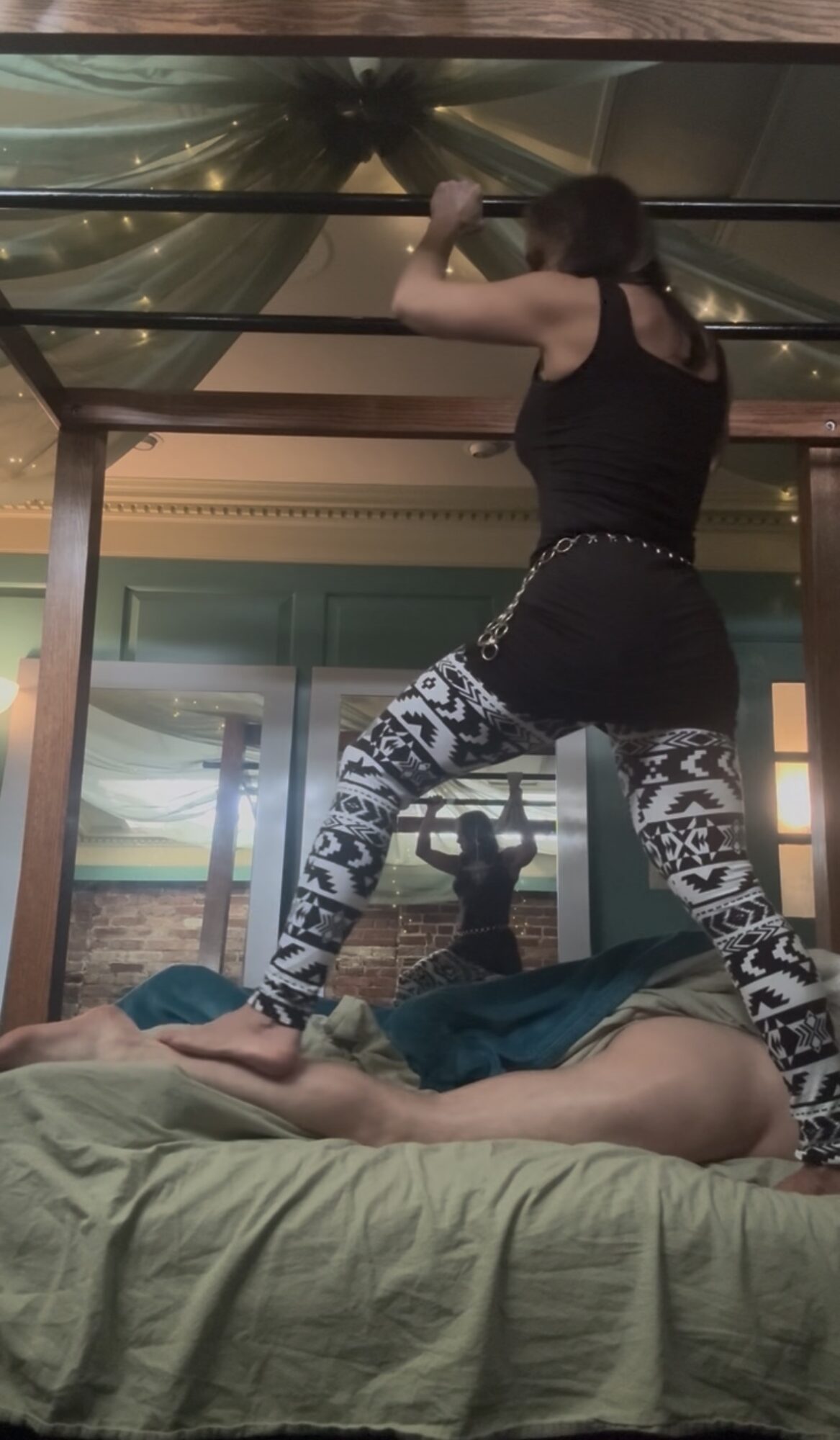 Person standing on bed, holding onto a metal bar, with a mirror reflecting their back and a person lying on the bed.
