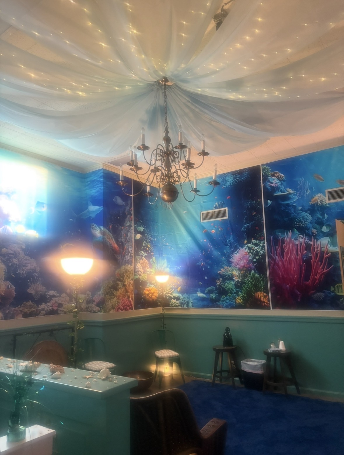 Room with underwater-themed mural, chandelier hanging from ceiling, and furniture including chairs and a table.