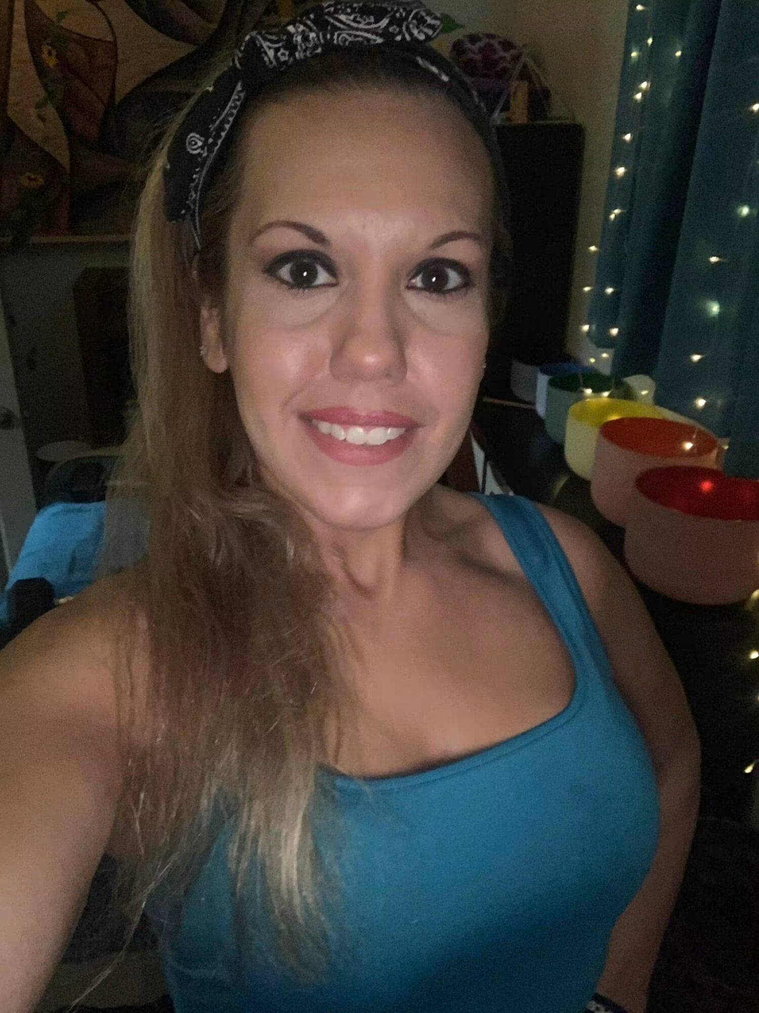 Smiling woman with long hair wearing a blue top, in a room with string lights and colorful bowls.